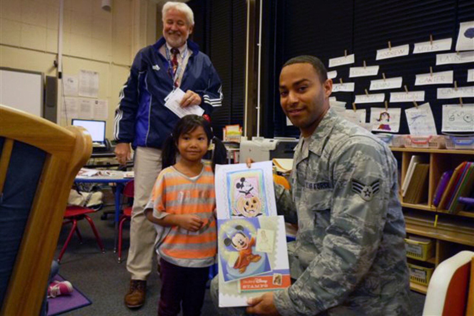 RAF ALCONBURY, United Kingdom – Senior Airman Jarron Webster, 423rd Communications Squadron Alconbury Post Office, presents Alysa Camacho with “The Art of Disney” stamp book after she was announced as one of nine winners of the 2013 October National Stamp Collecting Month coloring contest the Alconbury Post Office coordinated at the Alconbury Elementary School. (Photo courtesy of Linda Paine)