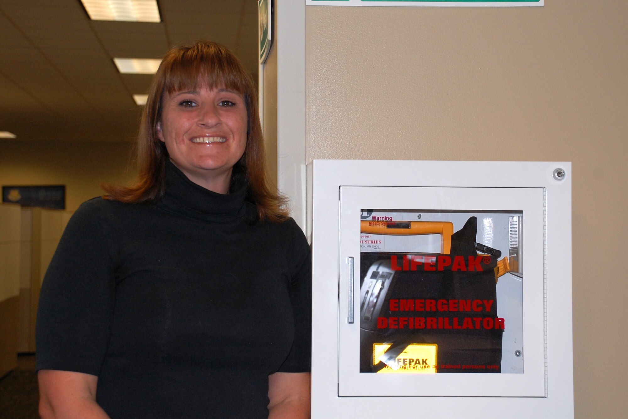 JOINT BASE SAN ANTONIO-LACKLAND, Texas -- Cassandra Price stands next to the automated external defibrillator, or AED, near her desk here. Her son Joshua is alive today thanks to quick responders and the use of an AED. Price is a member of the Air Force Civil Engineer Center's Installation Directorate Real Property Management office. (U.S. Air Force photo/Shannon Carabajal)