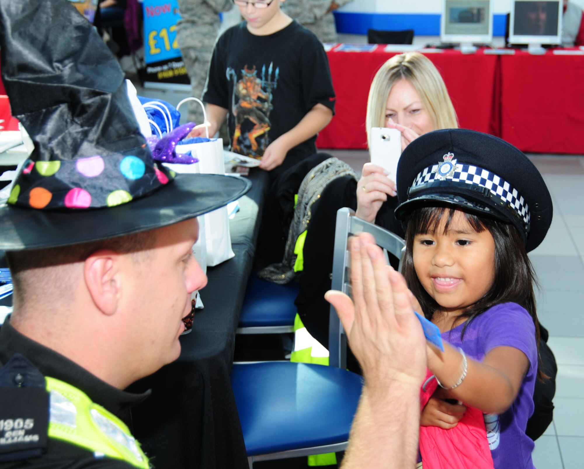 Police Constable Den Williams, left, Cambridgeshire Police from Ely, Cambridgeshire, trades hats with Myra Simmons, 4, as he chats to her mom, Aide Simmons from Albuquerque, N.M., about vehicle crime Oct. 25, 2013, at the BXtra on RAF Mildenhall, England, during the wrap-up event of Security Awareness Month. British police from Suffolk, Norfolk and Cambridgeshire, along with Ministry of Defence Police, and Airmen from the 100th Security Forces Squadron and 100th Air Refueling Wing Information Protection and Anti-Terrorism offices set up information booths and spoke with Team Mildenhall members. A military working dog display demonstrated how and why the MWDs are highly valuable assets to both the 100th SFS and RAF Mildenhall. (U.S. Air Force photo by Karen Abeyasekere/Released)