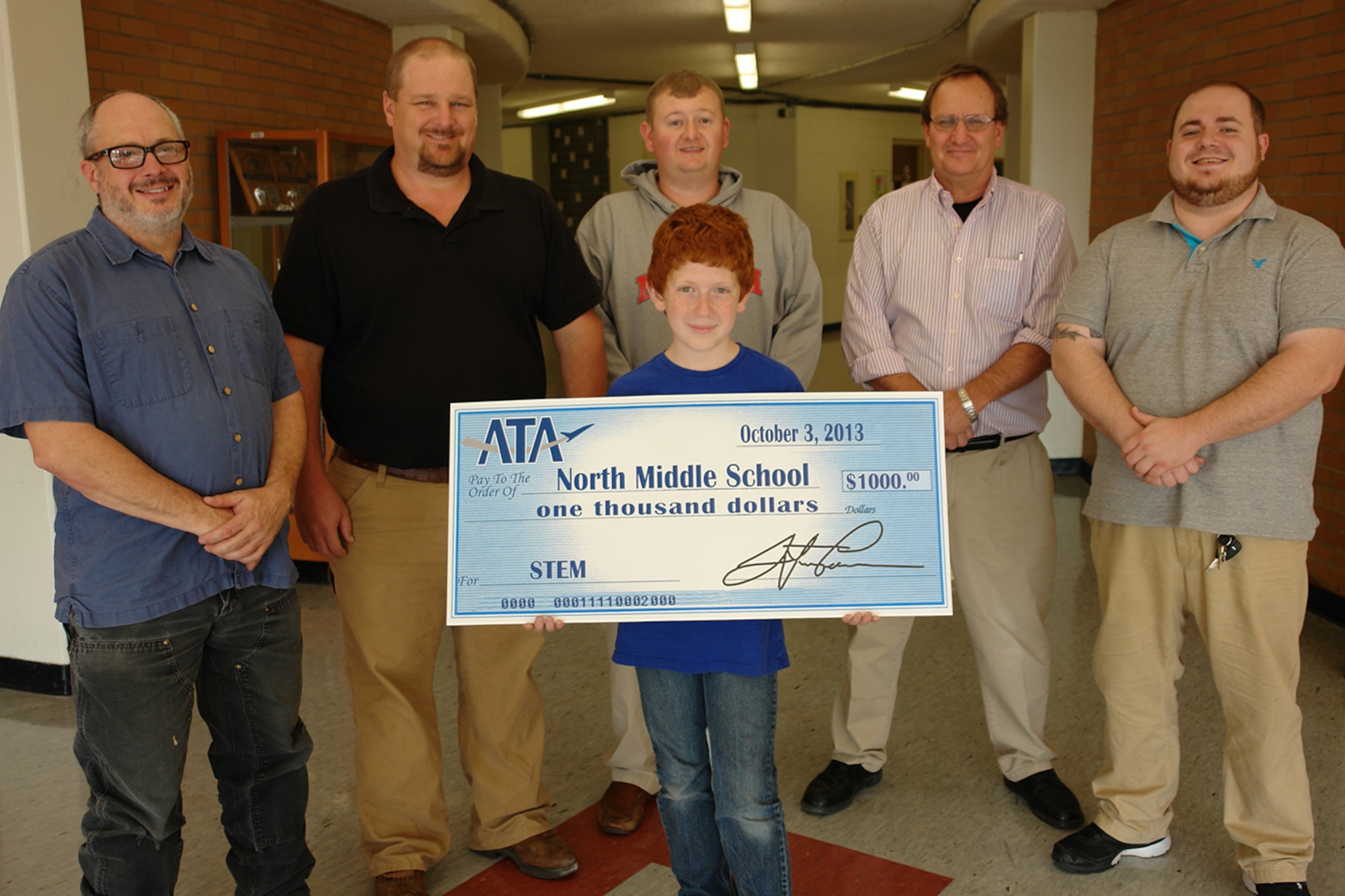 Two ATA employees make a check presentation to Jim Murphy, a math and science teacher at North Middle School.  Ricky Taylor (left), an ATA outside machinist and father of North Middle School student, Cameron Taylor (center); Jimmy Mitchell (2nd from left), an ATA test engineer, has a son, Mason, in Murphy’s math class; Avery Wiggs (center behind Cameron), social studies helper; Jim Murphy (4th from left) and Kyle Reid (right), another Tech Club coach who will help students learn programing language and computer code.