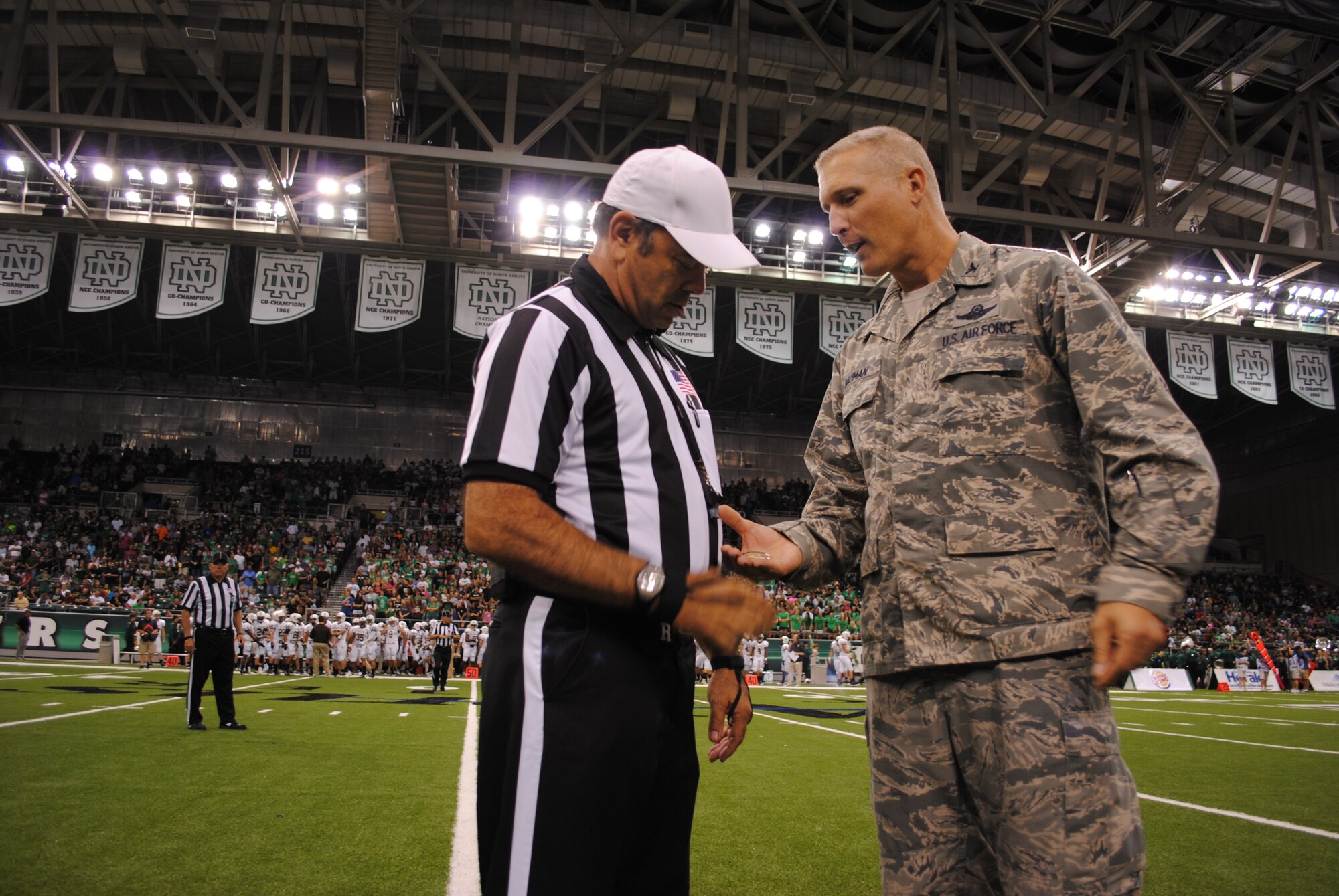 Col. Paul Bauman, 319th Air Base Wing commander, reciprocates NCAA football referee, Roger Stewart’s gift of the official game coin by giving him one of his wing commander’s coin, Aug. 29, 2013, at the Alerus Center in Grand Forks, N.D. Bauman was one of two honorary team captains for the University of North Dakota football game against Valparaiso University. Military members and their families were treated with a free tailgate party meal and free tickets to the game as part of the 16th Annual Maury Rothkopf Military Appreciation Day sponsored by the University of North Dakota  and the Grand Forks and East Grand Forks, Minn., Chambers of Commerce. (U.S. Air Force photo/Staff Sgt. Luis Loza Gutierrez) 
