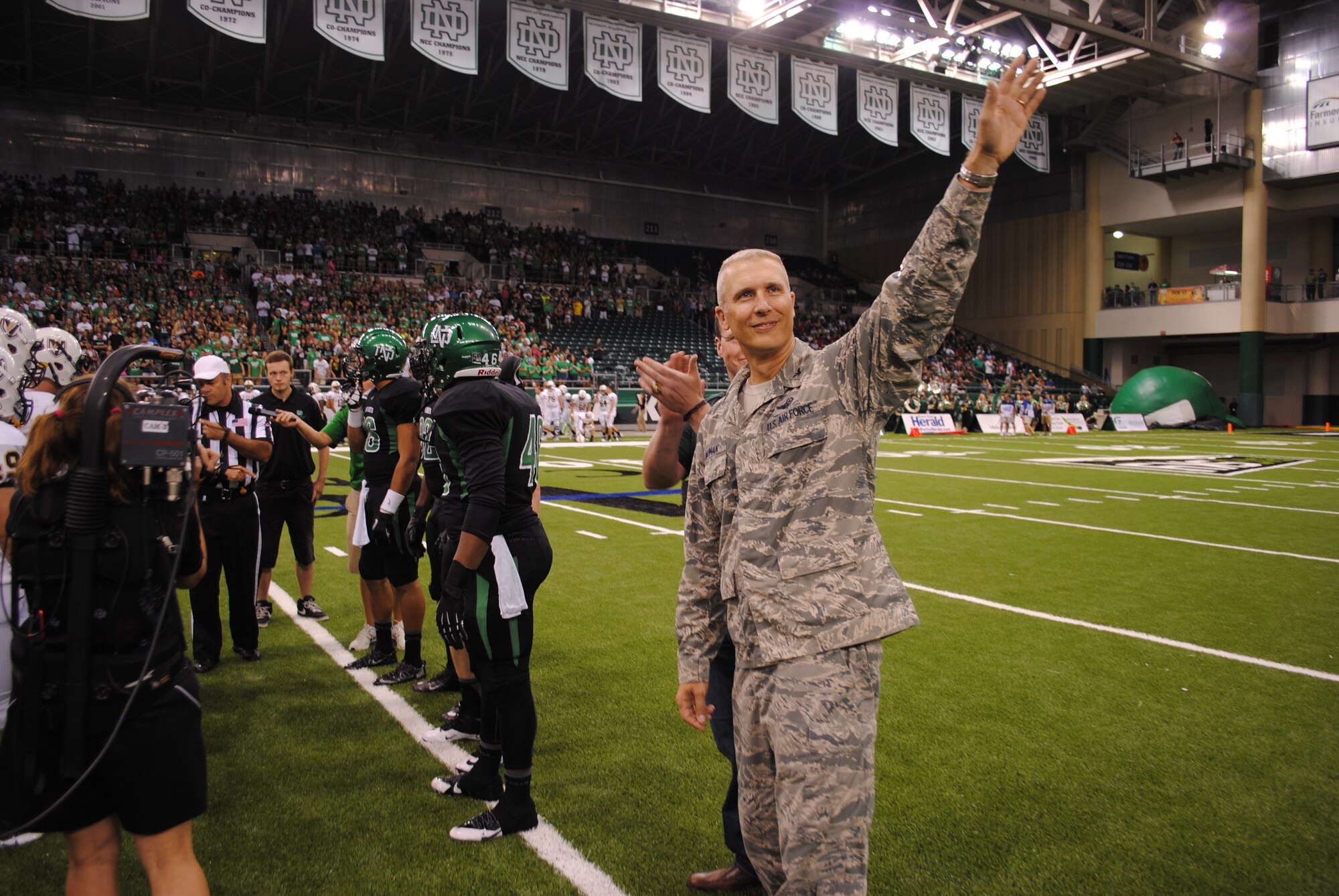 Col. Paul Bauman, 319th Air Base Wing commander, waves to the crowd from the center of the football field after being introduced as one of two honorary UND team captains for the football game against Valparaiso University, Aug. 29, 2013, at the Alerus Center in Grand Forks, N.D. Military members and their families were treated to a free tailgate party meal and free tickets to the game as part of the 16th Annual Maury Rothkopf Military Appreciation Day sponsored by the University of North Dakota  and the Grand Forks and East Grand Forks, Minn., Chambers of Commerce. (U.S. Air Force photo/Staff Sgt. Luis Loza Gutierrez) 
