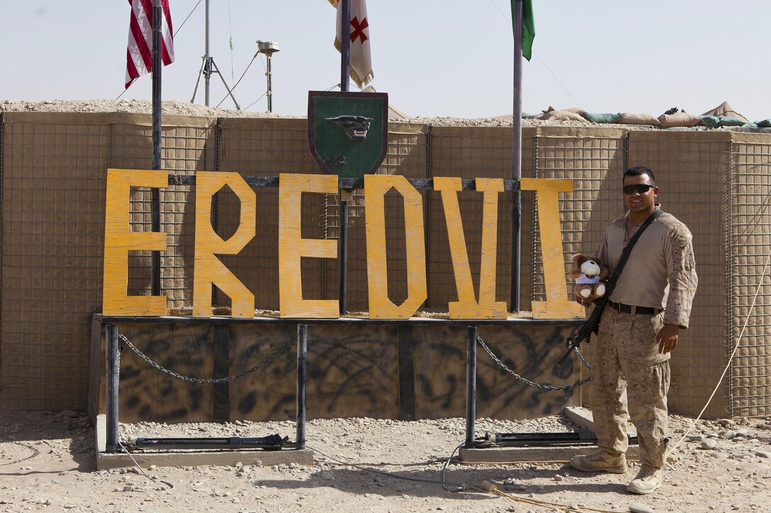Sgt. Anthony Ortiz, a California native and combat cameraman with Combat Logistics Regiment 2, Regional Command (Southwest), poses with a teddy bear at the sign for Combat Outpost Eredvi, Helmand province, Afghanistan, Aug. 13, 2013. Ortiz took the doll with him during his missions in the province before sending it home to his daughter for her third birthday.