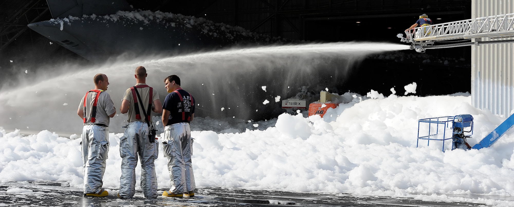 Mr. Barry Williams, a civilian fire fighter with the 436th Civil Engineer Squadron, Fire Department, sprays water on fire suppression foam  in hangar 706 while other firefighters watch on Sept. 16, 2013 at Dover Air Force Base, Del. Personnel from the Dover Fire Department responded to the inadvertent release of fire suppression foam and then worked to dissipate the foam using water sprayed from a ladder truck so clean-up efforts could begin. (U.S. Air Force photo/Greg L. Davis)