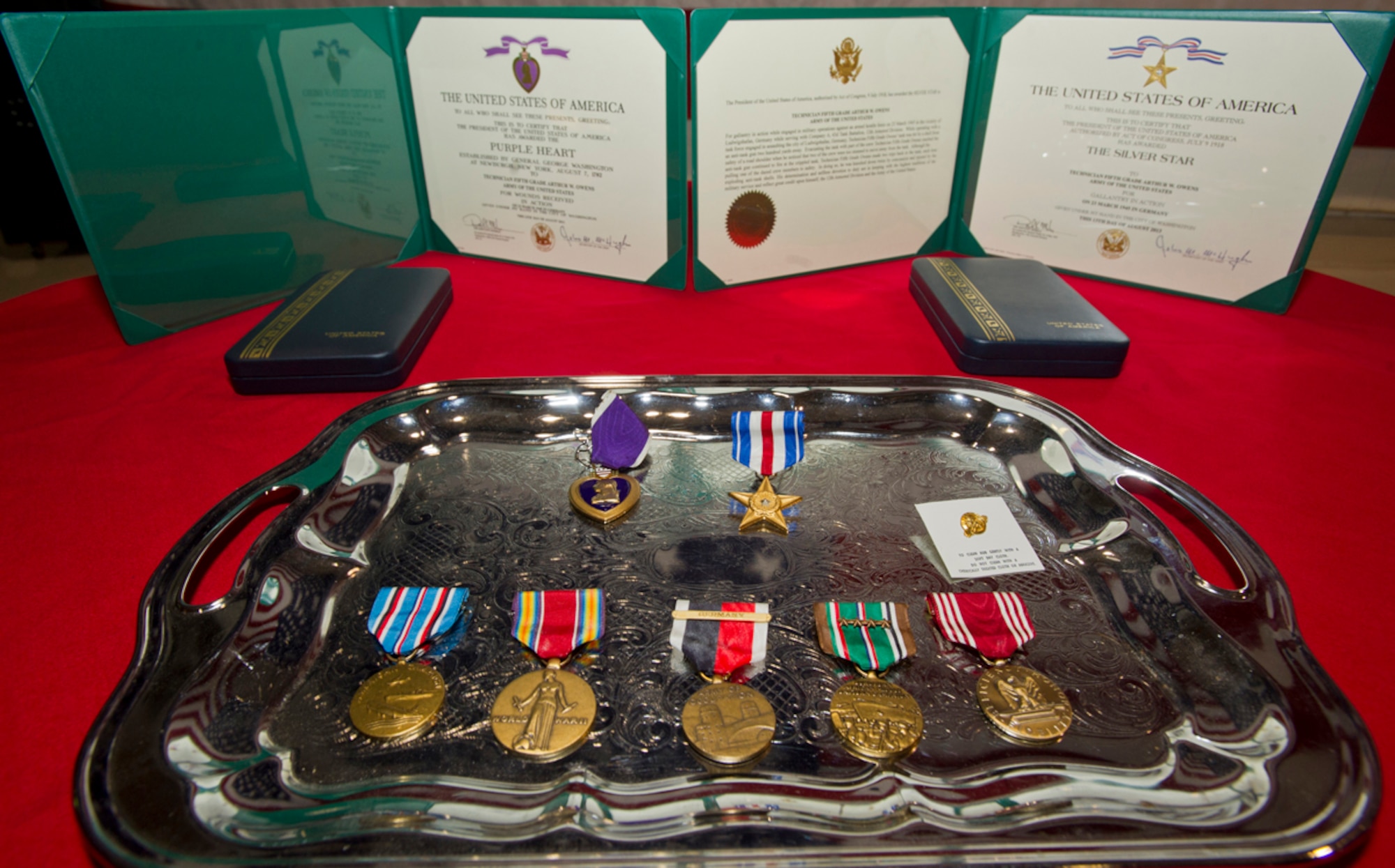 (From center-left) A Purple Heart, Silver Star, American Campaign Medal, WWII Victory Medal, WWII Occupation Medal with "Germany" clasp, European/African/and Middle Eastern Campaign Medal with four bronze stars, and a Good Conduct Medal, lie on a table before World War II veteran Arthur W. Owens is awarded the Silver Star and Purple Heart 68 years after his heroic actions while fighting near Ludwigshafen, Germany. On March 23, 1945, Owens' tank was hit by a shell from an anti-tank gun.  After evacuating the vehicle, he returned twice under heavy enemy fire -- and despite his wounds -- to rescue injured comrades. After a review of his records, Secretary of the Army John McHugh recently signed orders to recognize Owens. Brig. Gen. Mike Bridges, commander of the Alaska Army National Guard, presented the medals during a ceremony at the Alaska National Guard headquarters on Joint Base Elmendorf-Richardson, Thursday, Sept. 19, 2013. (U.S. Air Force photo by Justin Connaher/Released)