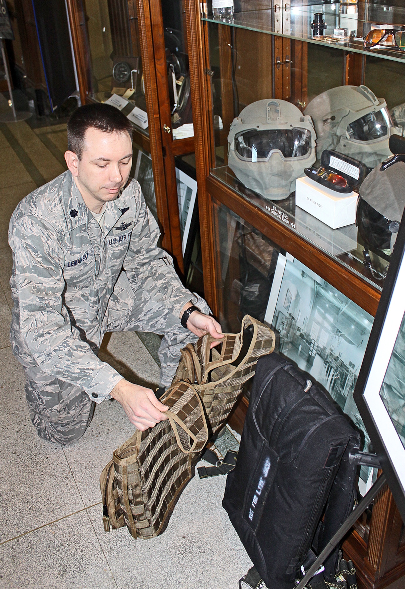 Lt. Col. Chris Lemanski kneels beside a low profile parachute and an aircrew survival vest and beneath a glass-encased helmet and aviator lenses all developed under the auspices of the Agile Combat Support division.