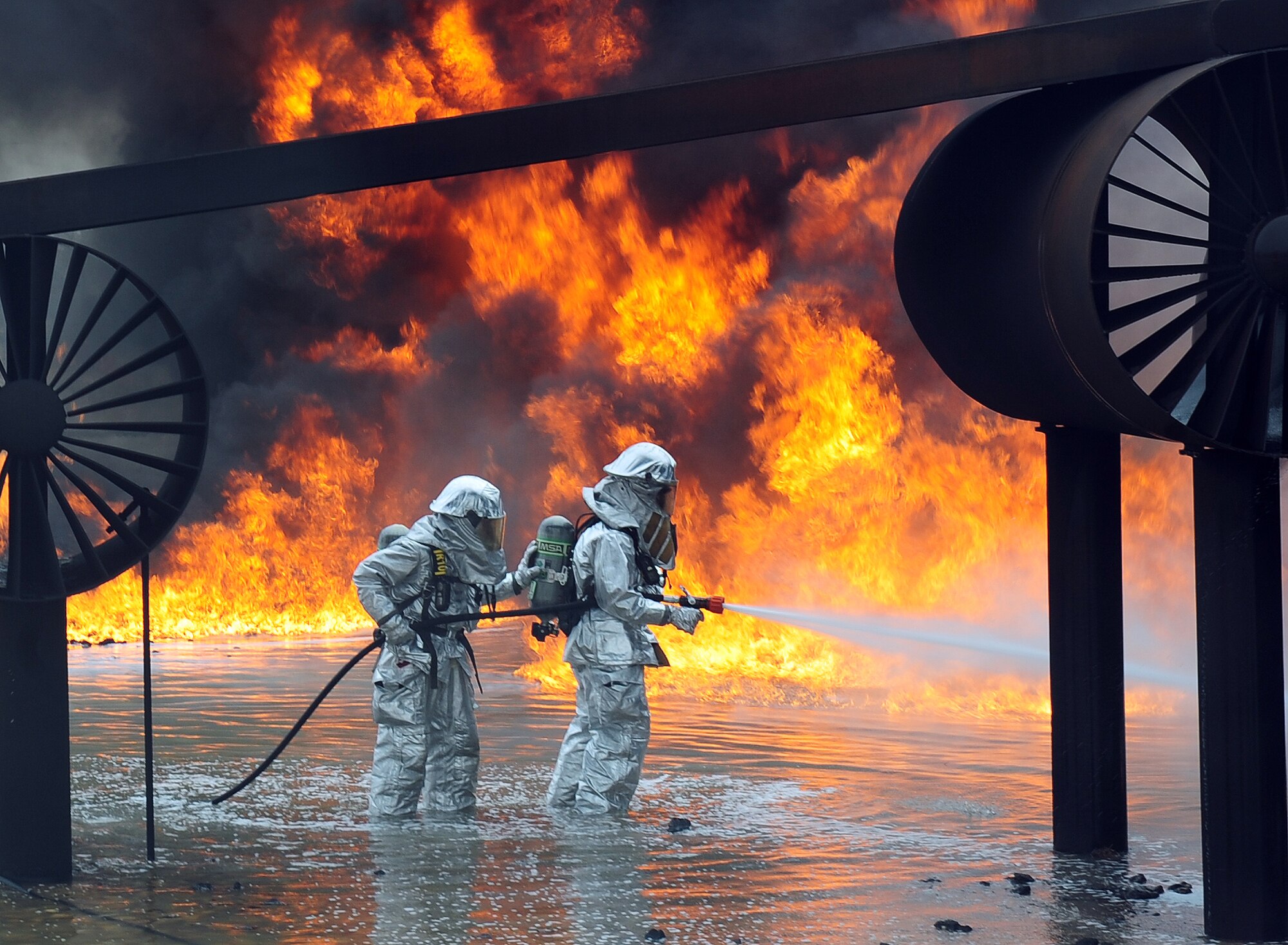 U.S. Air Force firefighters from the 7th Civil Engineer Squadron spray water onto a fire during a live fire training exercise April 2, 2014, at Dyess Air Force Base, Texas. In addition to wearing their fire proximity suits, firefighters must also wear self-contained breathing apparatus’ that provide a source of clean oxygen, enabling them to work in the presence of smoke or other super-heated gases.  (U.S. Air Force photo by Senior Airman Kia Atkins/Released)