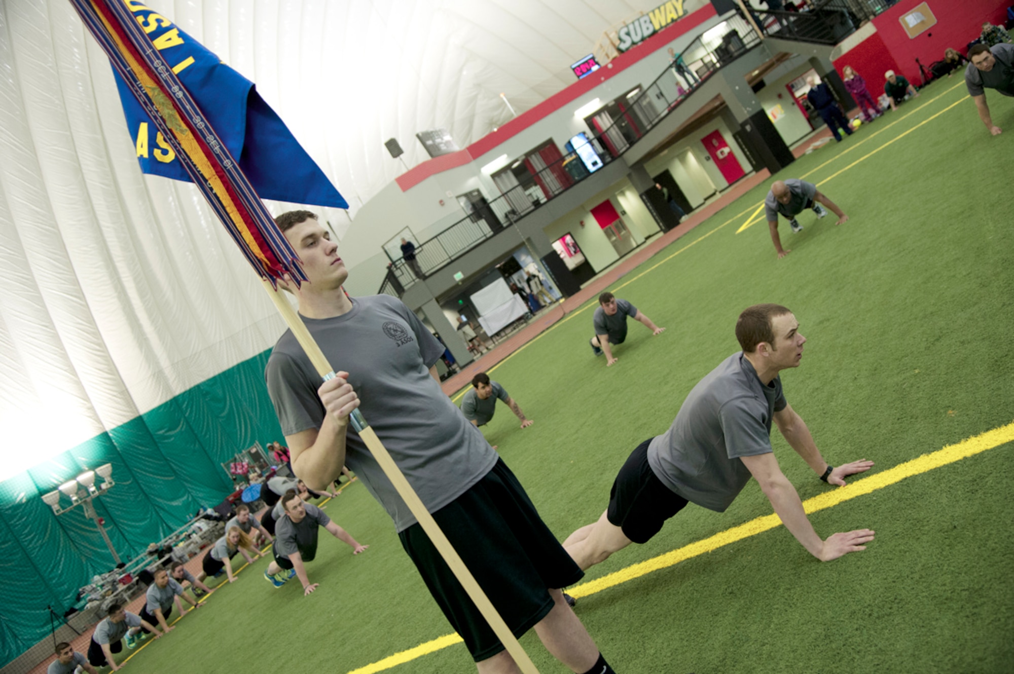 Airman 1st Class Jesse Rehkop holds the 3rd Air Support Operations Squadron guidon while Airman 1st Class Trenton Collins leads the Tactical Air Control Party’s memorial push-up event after the annual 24-hour memorial run inside the Alaska Dome in Anchorage Friday. (U.S. Air Force photos/Staff Sgt. Sheila deVera)