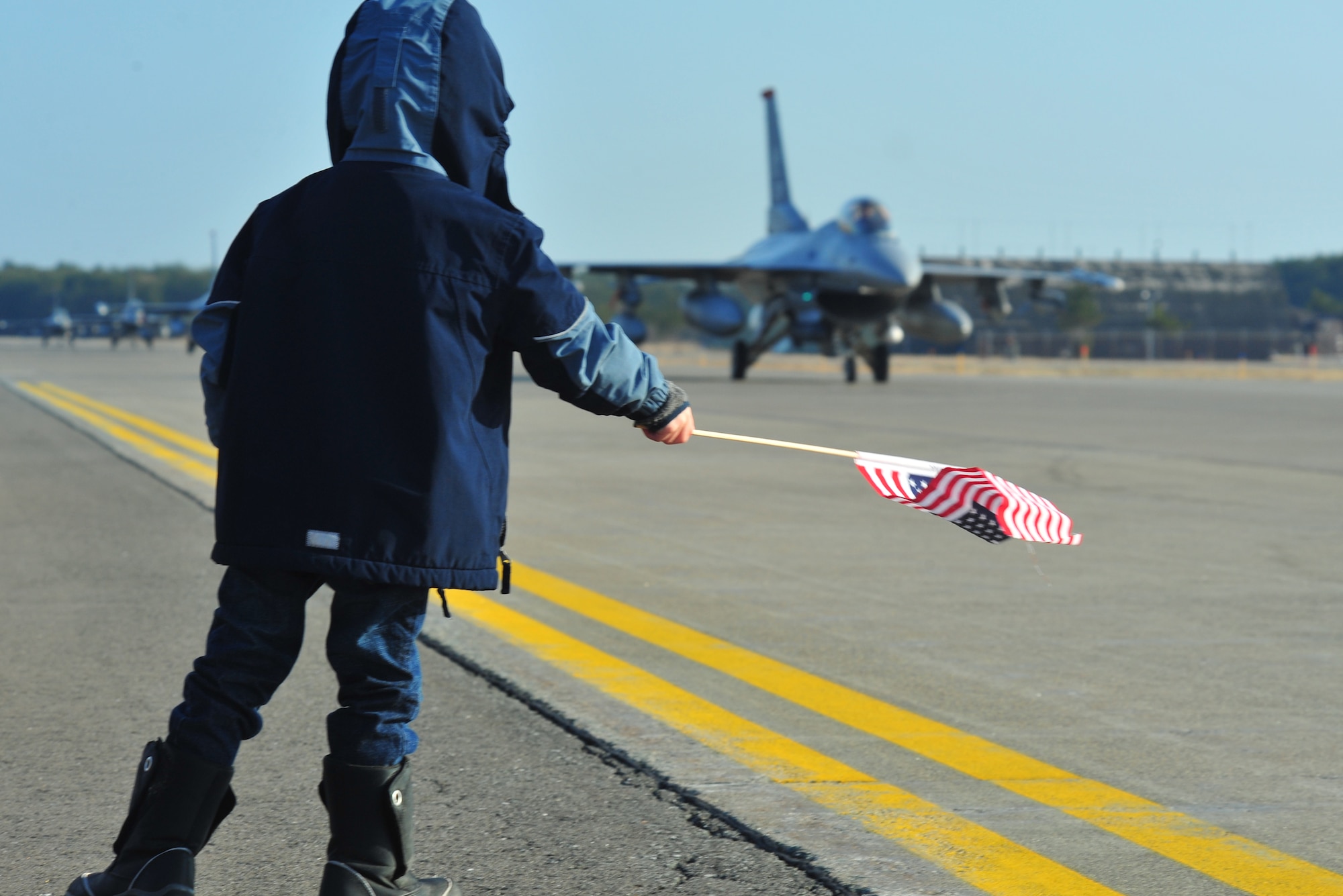 The son of a 13th Fighter Squadron pilot waves an American flag at a fleet of oncoming F-16 Fighting Falcons at Misawa Air Base, Japan, April 8, 2014. Fighter jets from the 13 FS revved up their engines early morning as they readied for deployment in support of combatant commanders down range. (U.S. Air Force photo/Senior Airman Jose L. Hernandez-Domitilo)
