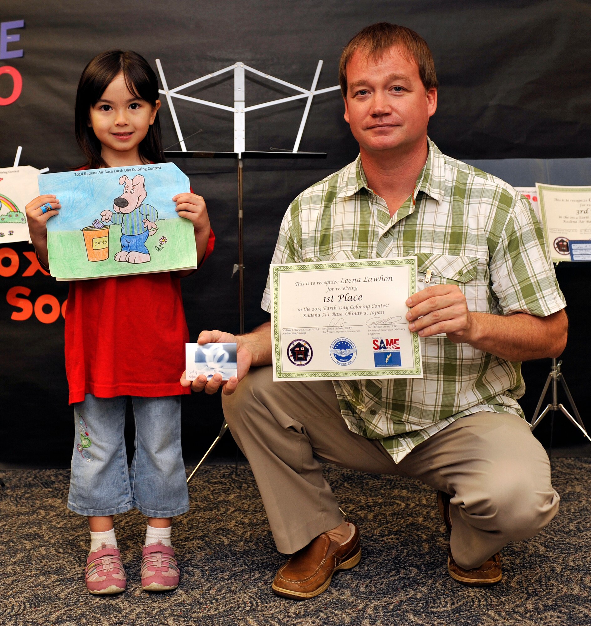 Leena Lawhon, 6, poses for a photo while holding her art piece with Vincent Adams, 718th Civil Engineer Squadron solid waste manager, during the Earth Day art award ceremony at Bob Hope Primary School on Kadena Air Base, Japan, April 23, 2014. There were a total of 49 entries for the coloring contest and 27 entries for the poster. The prizes for the top three winners were base exchange gift cards: $100 for first, $50 for second, and $25 for third place winners. Leena became the first place winner in the coloring contest. (U.S. Air Force photo by Naoto Anazawa)