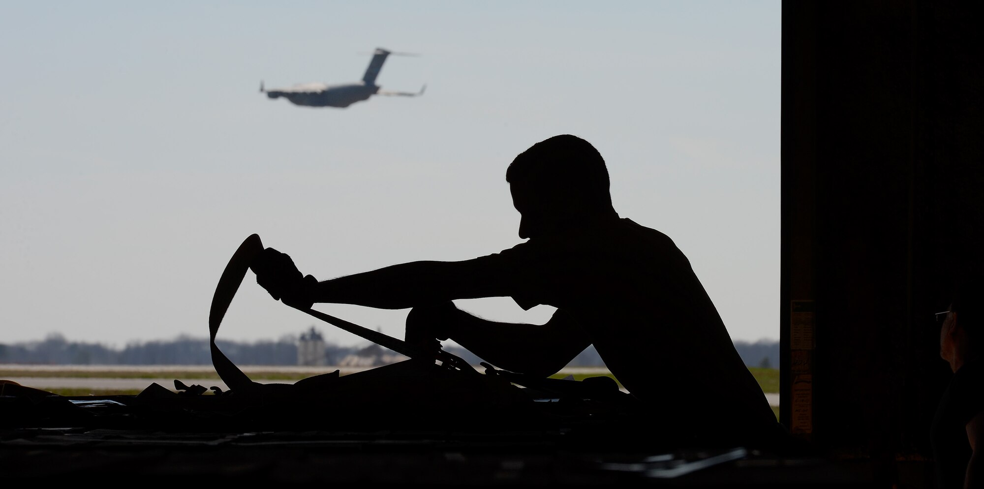 Tech. Sgt. Paul Osborn, 436th Aerial Port Squadron NCO in charge of in-transit munitions section, pulls a cargo strap tight as he configures cargo on April 10, 2014, at Dover Air Force Base, Del. Most of the munitions processed by the section are transported via airlift aircraft such as the C-17A Globemaster III captured in-flight in the background. (U.S. Air Force photo/Greg L. Davis) 
