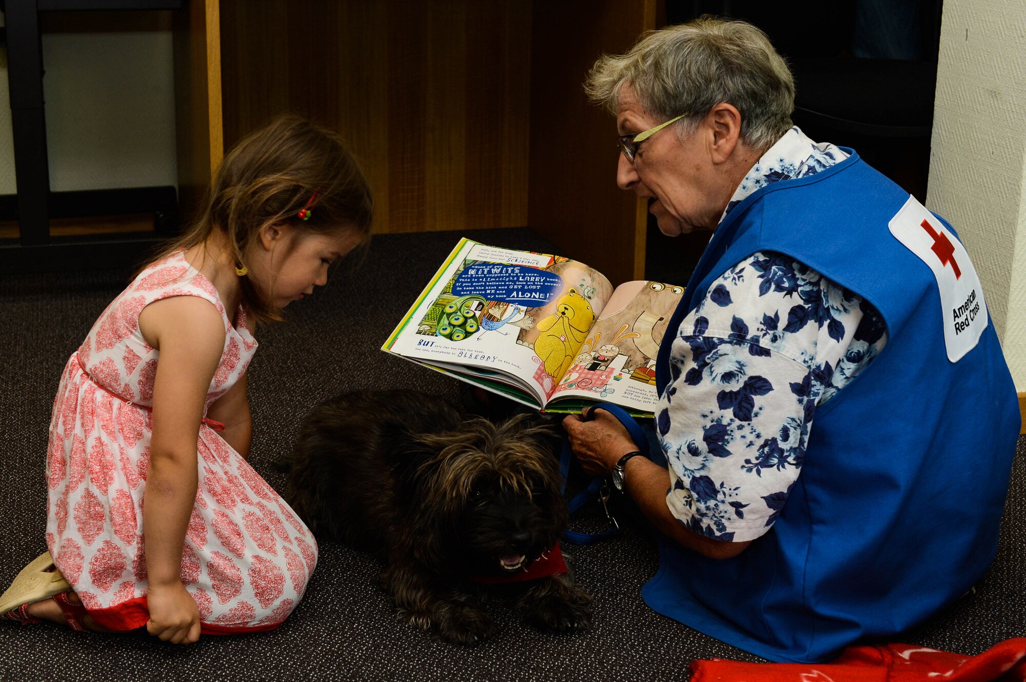 Lucia Padillabarg (left) and Anka Jessie (right), read a book to Pepper, a Red Cross Morale Dog, in the library at Spangdahlem Air Base, Germany, July 31, 2014.  Pepper helps the Red Cross by meeting with patients at hospitals like Landstuhl Regional Medical Center in an effort to reduce stress and raise morale. (U.S. Air Force photo by Airman 1st Class Kyle Gese/Released)