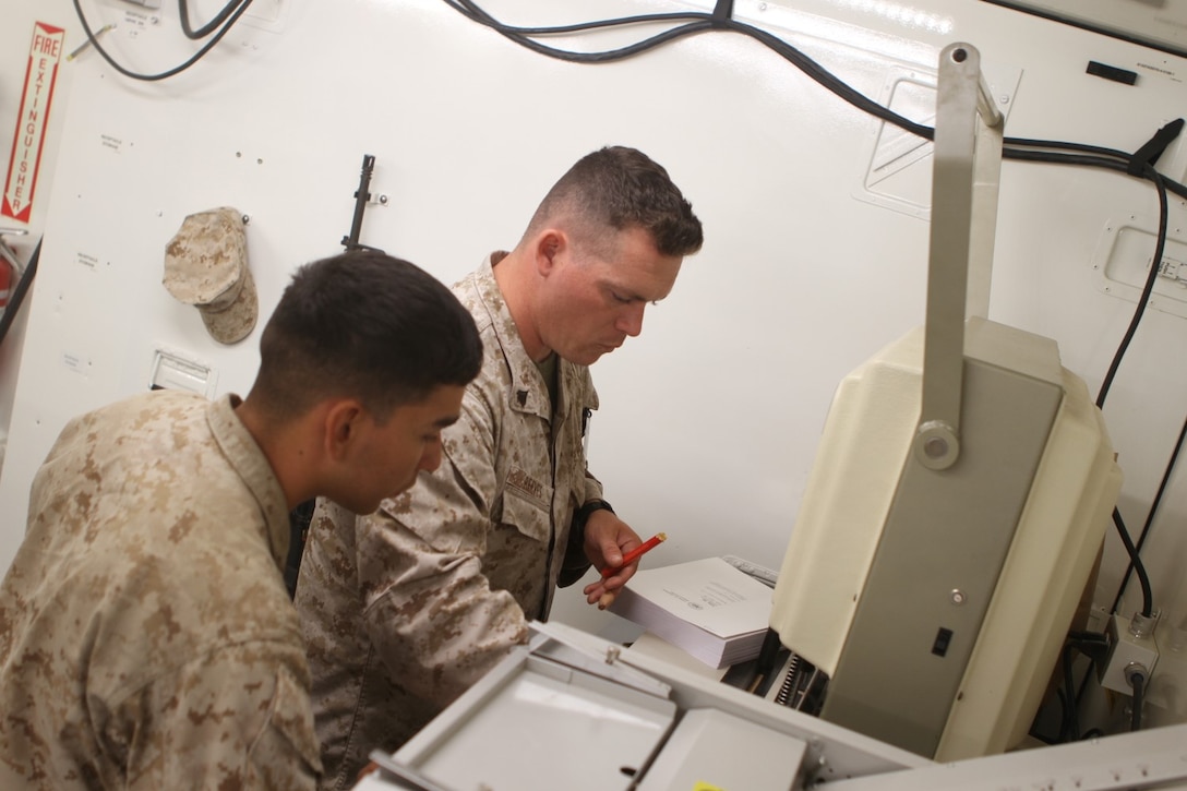 Cpl. Robert Reeves, combat camera reproduction chief with 1st Marine Expeditionary Brigade, guides Lance Cpl. James A. Trevino, a combat videographer also with 1st MEB, on how to operate machines for reproduction and printing jobs inside a Tactical Imagery Production System during Large Scale Exercise 2014 aboard Marine Corps Air Ground Combat Center Twentynine Palms, Calif., Aug. 2, 2014. LSE-14 is a bilateral training exercise being conducted by 1st MEB to build U.S. and Canadian forces' joint capabilities through live, simulated, and constructive military training activities from Aug. 8-14. (U.S. Marine Corps photo by Lance Cpl. Angel Serna/Released)