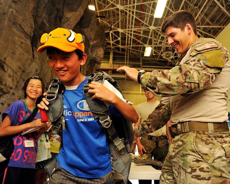 U. S. Air Force Senior Airman Austen Carroll, 31st Rescue Squadron pararescueman, helps Shinji Kamemura, put a parachute on during a visit on Kadena Air Base, Japan, Aug. 4, 2014. Thirteen primary school children from Achi Village, Nagano, and five primary school children from Okinawa City visited Kadena as part of an annual Okinawa visit to the base. Japanese primary school children visit during this time every year to learn about significant pieces of history and engage with American children their age during the visit here. (U.S. Air Force photo by Naoto Anazawa/Released)