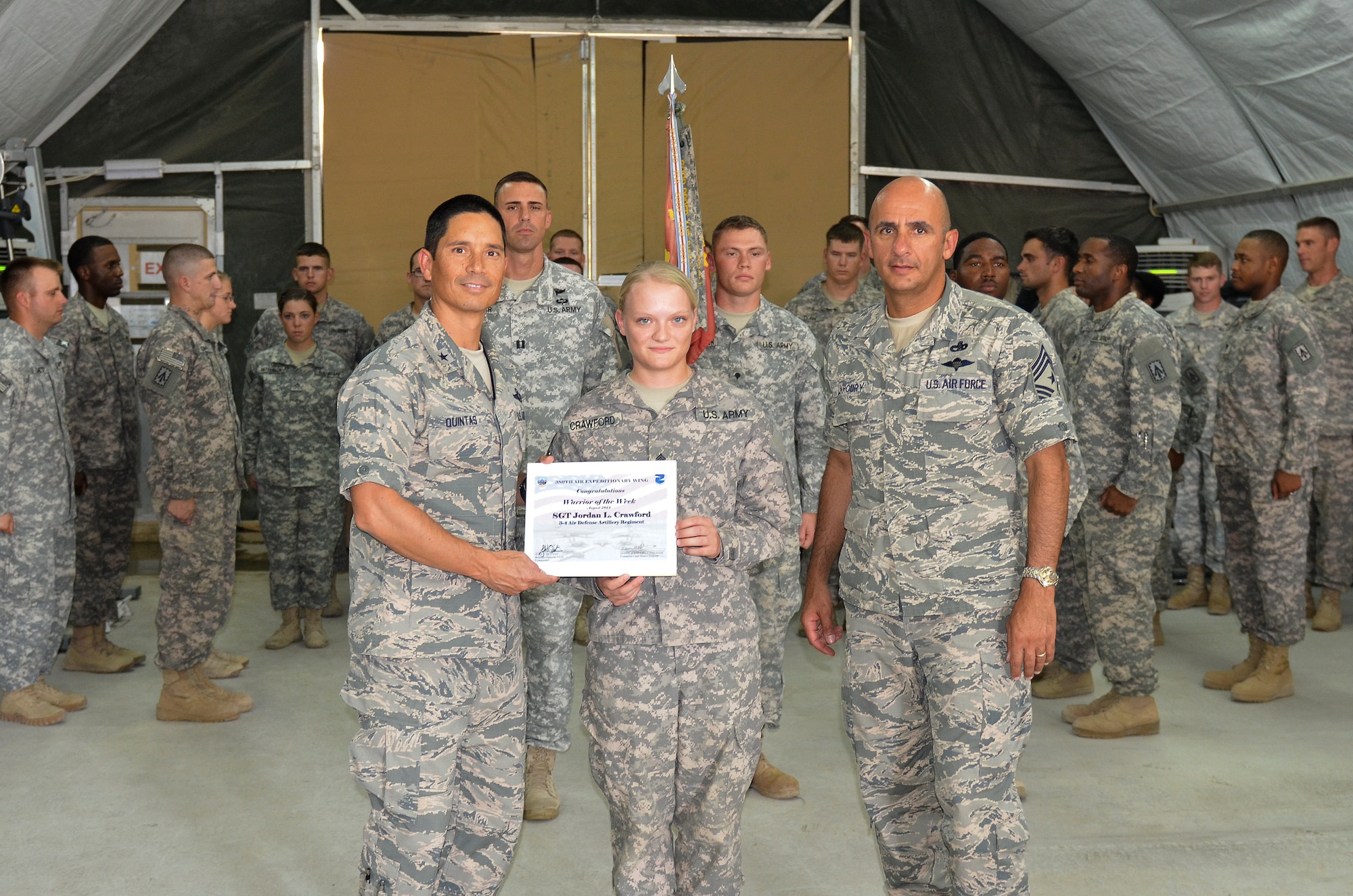 Air Force Brig. Gen. John Quintas, 380th Air Expeditionary Wing commander, and Chief Master Sgt. Samer Alkhoury, 380th Air Expeditionary Wing command chief, present Army Sgt. Jordan Crawford, Bravo Battery, 3-4 Air Defense Artillery Regiment, a warrior of the week certificate at an undisclosed location in Southwest Asia Aug. 6, 2014. Crawford is deployed from Fort Bragg, N.C. 