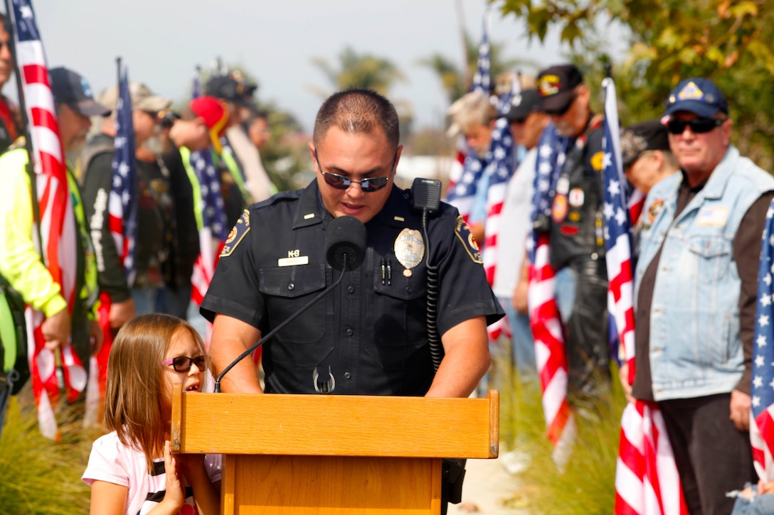 2nd Lt. Benjamin Maple, the kennel master with the Marine Corps Police Department Camp Pendleton K-9 Unit, gives a speech at a memorial dedication with his daughter Taylor Star Maple, 6 at the Pacific Views Event Center’s Memorial Garden here, Aug. 9.  The Dawgs Project, a company dedicated to military working dog handlers of all services, unveiled the memorial to commemorate K-9 handling service members.