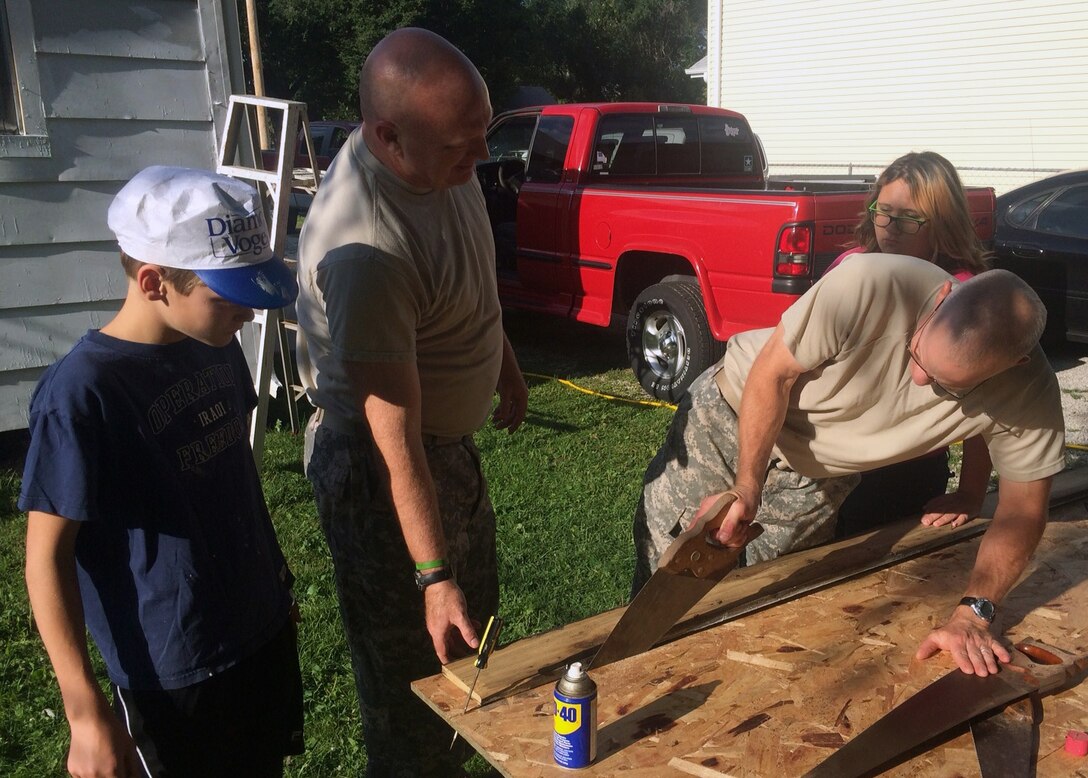 Col Joel Cross, Omaha District Commander saws the plywood used to replace the old cellar door of the home being painted for Paint-A-Thon 2014, while Lt Col Michael Sexton, Deputy Commander, oversees the process. Meanwhile Brian Cross and a teenage neighbor girl look on. 