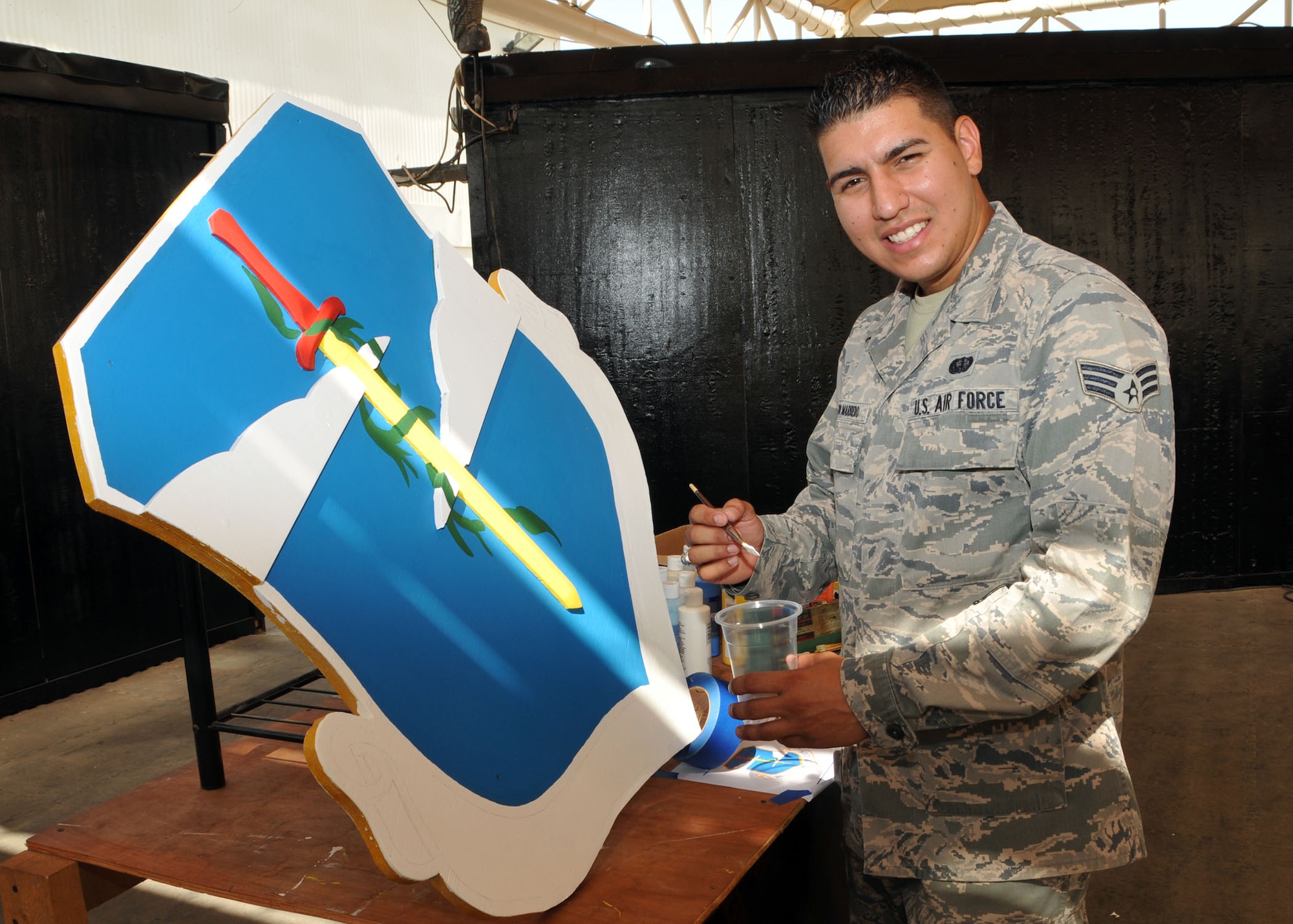Air Force Senior Airman Bernabe Negron puts lettering on a three-dimensional 380th Air Expeditionary Wing shield he painted at the base at an undisclosed location in Southwest Asia. Negron created the emblems and shields used on the walls of the wing’s Community Activity Center pavilion stage. (U.S. Air National Guard photo by Senior Master Sgt. Eric Peterson/Released)