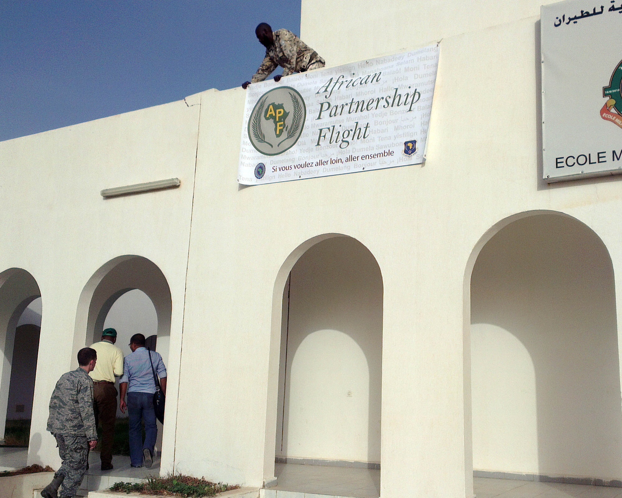 U.S. Air Force Airmen from U.S. Air Forces in Europe and Air Mobility Command enter the main training building before opening the opening ceremony of the African Partnership Flight Mauritania in Atar, Mauritania while a Mauritanian Airmen hangs an APF banner on the building Aug. 30, 2014. APF is multi-national week-long training event that is designed to give African partners skills on ground and flight safety, Intelligence Surveillance Reconnaissance, and command operations that will build aviation capacity, enhance regional cooperation and increase interoperability throughout the region. (U.S. Air Force photo/Master Sgt. Brian Boisvert/Released)