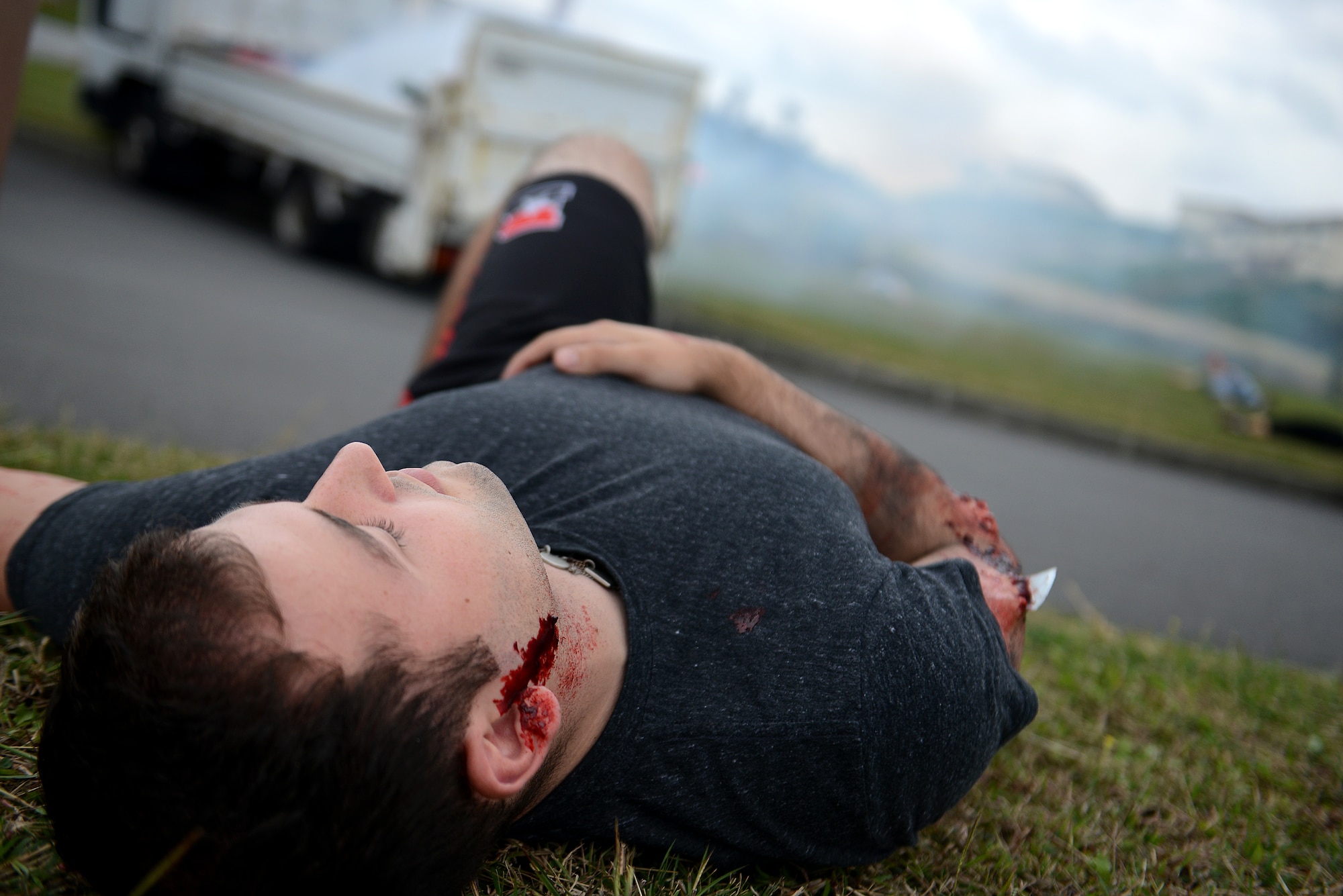 A victim of a simulated hazardous material vehicle crash lies in the grass near a smoking vehicle during a mission focused exercise on Kadena Air Base, Japan, Dec. 2, 2014. During MFEs, wing inspection team members use special makeup on simulated casualties to provide realism to the different training scenarios. (U.S. Air Force photo by Senior Airman Maeson L. Elleman/Released) 