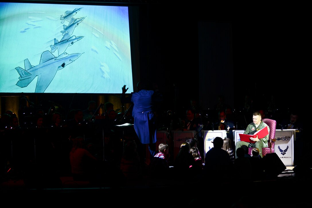 U.S. Air Force Maj. Robert Burgon, 27th Fighter Squadron assistant director of operations and pilot, reads a book titled “Robbie the Raptor,” while the U.S. Air Force Heritage of America Band performs a musical adaptation of the story at their holiday concert, “Through Children’s Eyes,” at Jamestown High School in Williamsburg, Va., Dec. 2, 2014. During the reading, children from the crowd were encouraged to join Burgon on stage. (U.S. Air Force photo by Senior Airman Kimberly Nagle/Released)