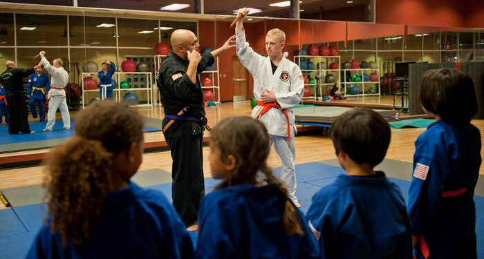 Smaller students look on as Renshi DeAndre Scott (black gi) instructs students to avoid knife attacks during the Nesei Bujutsu class at the Warrior Fitness Center on Nellis Air Force Base, Nev., Dec. 9, 2014. Students of all sizes are taught to disarm and subdue attacker using holds and pressure points.(U.S. Air Force photo by Staff Sgt. Victoria Sneed)