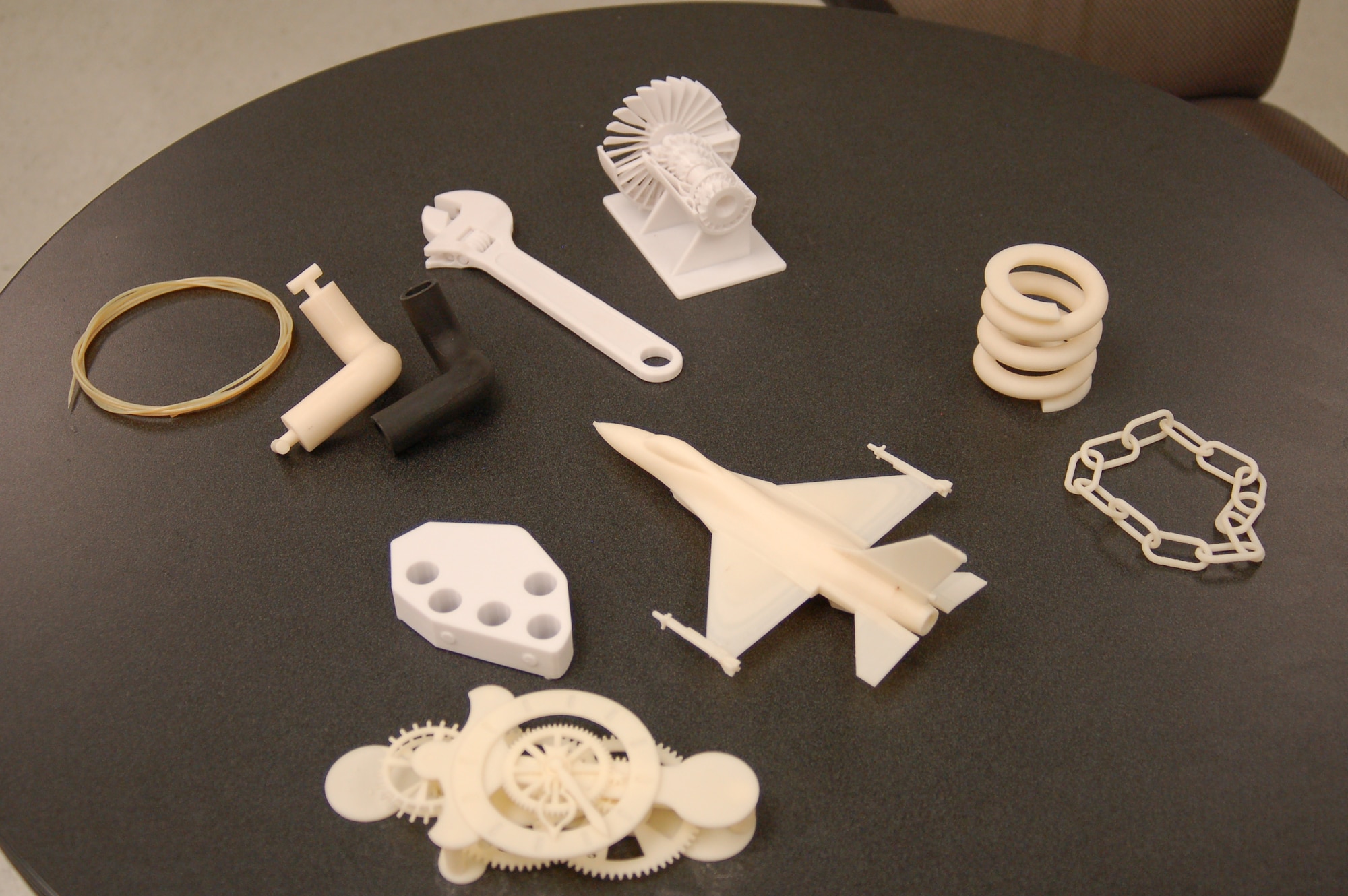 A thin line of thermoplastic, coiled at left, is used in a 3-D printer to create objects like those shown on the table as well as prototype parts for aircraft. (Air Force photo by Bill Orndorff)