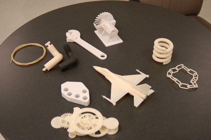 A thin line of thermoplastic, coiled at left, is used in a 3-D printer to create objects like those shown on the table as well as prototype parts for aircraft. (Air Force photo by Bill Orndorff)