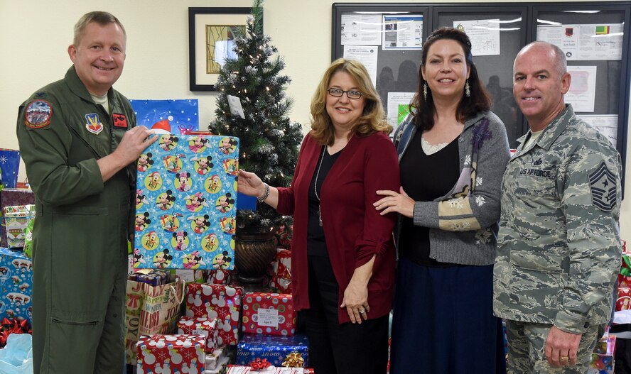 Col. James Cluff, 432nd Wing/432nd Air Expeditionary Wing commander, left, and Chief Master Sergeant Butch Brien, 432nd Wing/432nd AEW command chief, give the first of 300 presents to Indian Springs school representatives Kimberly Guerino and Kelly Miles during the 2014 Holiday Gift Drive open house at the Airmen Ministry Center at Creech Air Force Base, Nevada, Dec. 16, 2014. The Creech Chapel first began the gift drive in 2010, and this year’s donations supported 110 children who attend Indian Springs elementary, middle and high schools. (U.S. Air Force Photo by Tech. Sgt. Shad Eidson/released)