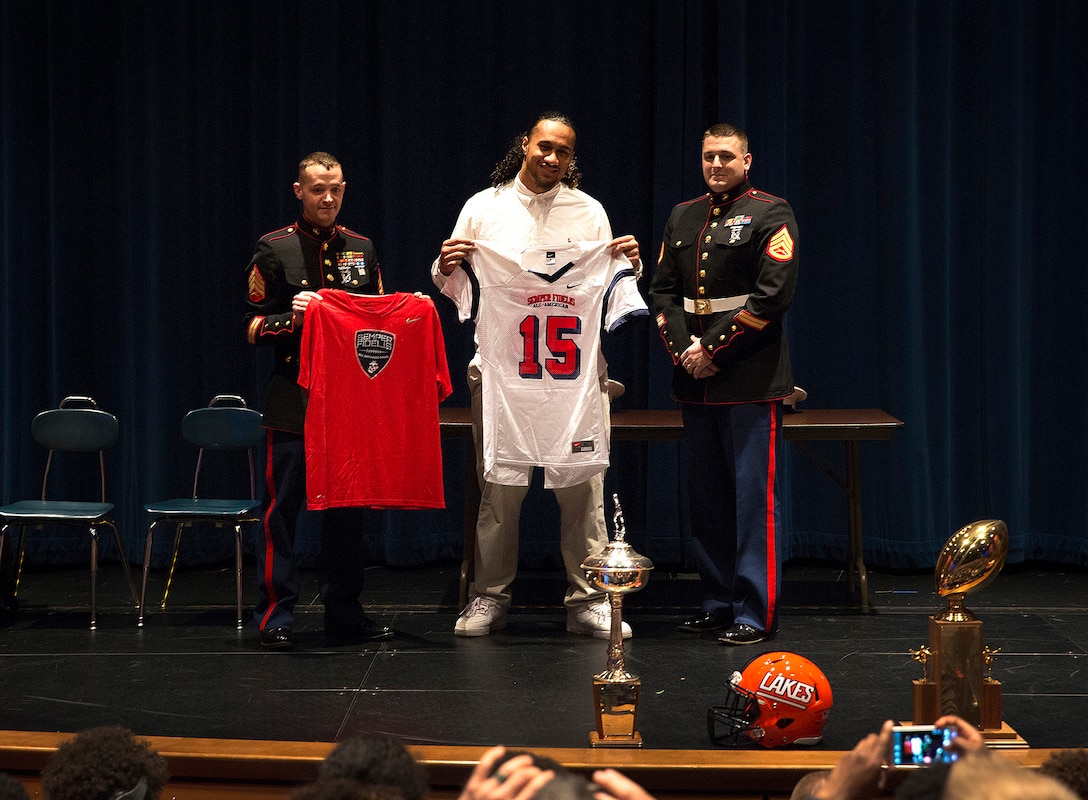 Benning Potoae (center), a defensive end for the Lakes High School Lancers varsity football team and 18-year-old native of Dupont, Washington, receives his 2015 Semper Fidelis All-American Bowl jersey from Sgt. Daniel Pluth (left), a Tacoma recruiter, and Staff Sgt. Cody Branham (right), the recruiter in charge of Recruiting Substation Puyallup, during a ceremony at Lakes High School in Lakewood, Washington, Dec. 16, 2014. Potoae is among approximately 100 student athletes from across the nation selected to participate in the game at the StubHub Center in Carson, Calif., Jan. 4, 2015. He joins cornerback Austin Joyner from Marysville-Pilchuck High School and offensive tackle Shane Lemieux from Yakima’s West Valley High School as the Washington State players selected for the 2015 game. (U.S. Marine Corps photo by Sgt. Reece Lodder)