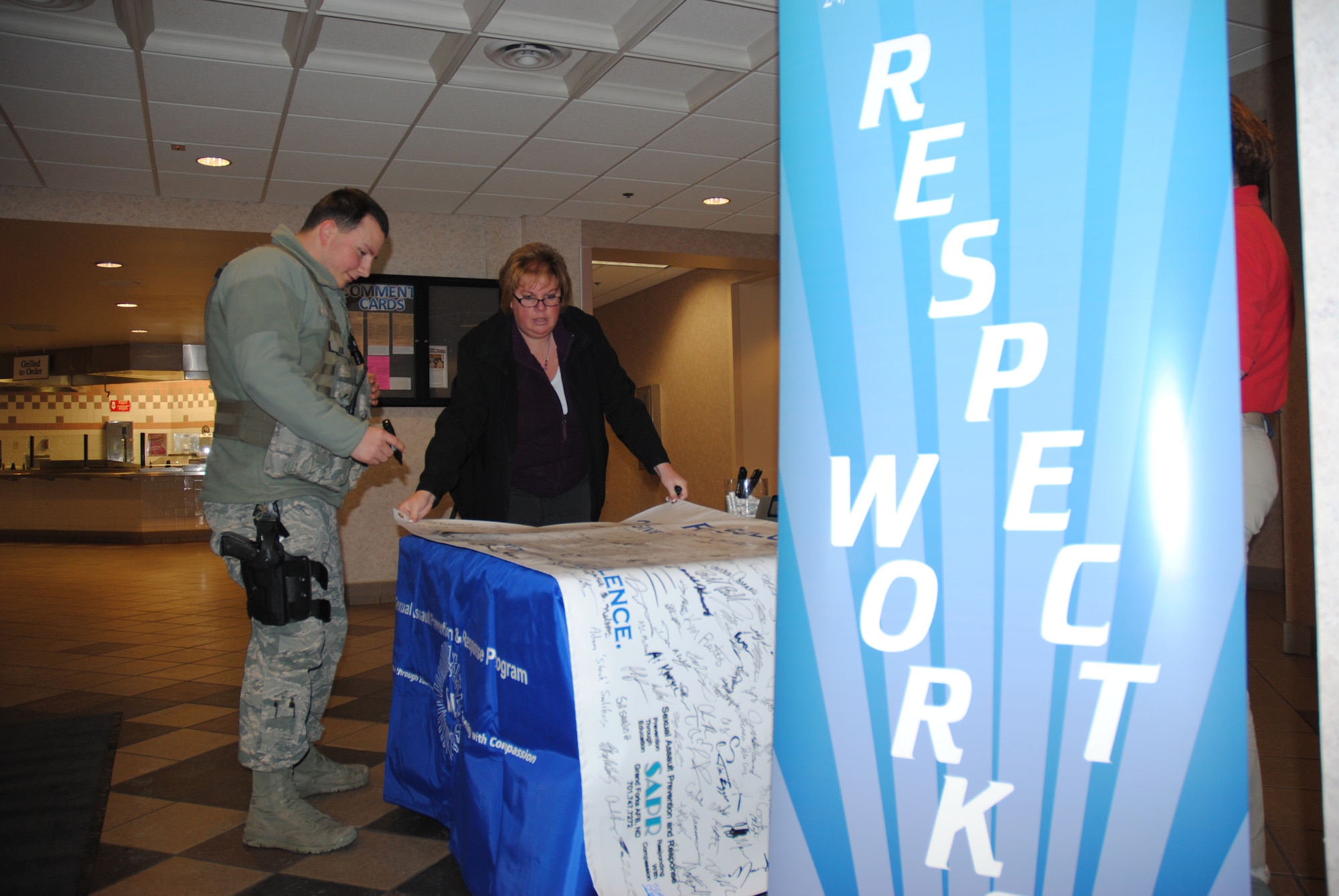 Sue Grollimund, 319th Air Base Wing Sexual Assault Prevention and Response Program assistant, helps Airman 1st Class Duane Alex, 319th Security Forces Squadron find a blank spot to sign his name to the 2015 Year of Respect banner, Dec. 18, 2014, at the Airey Dining Facility on Grand Forks Air Force Base, N.D.  From December 15 through19, the SAPR office took its “Resolution: Respect” campaign out to various base facilities. (U.S. Air Force photo/Staff Sgt. Luis Loza Gutierrez)