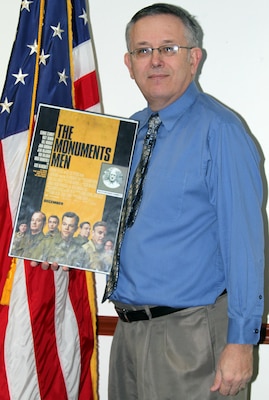 Little Rock District Deputy District Counsel Ralph Allen stands with a movie poster for “The Monuments Men,” a current film about the dangerous job his grandfather, Ralph W. Hammett, held during World War II. 
