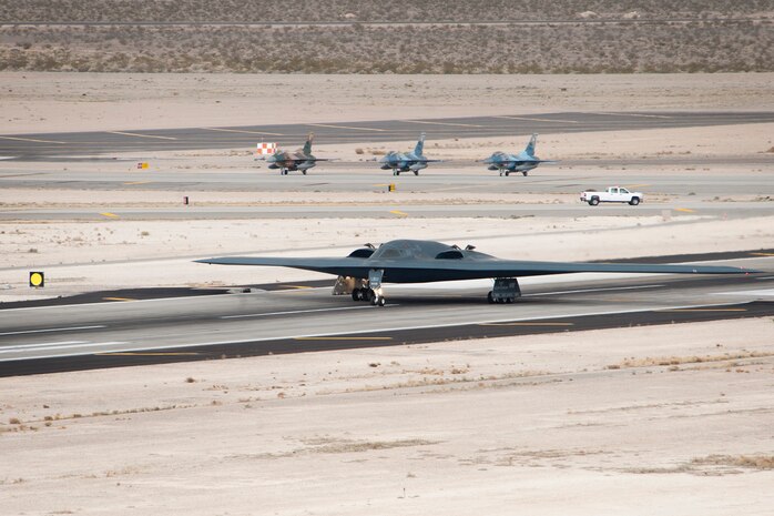 Three F-16 Fighting Falcons assigned to the 64th Aggressor Squadron hold short of the runway as a B-2 Spirit, assigned to the 13th Bomb Squadron from Whiteman Air Force Base, Mo. takes off during Red Flag 14-1, Feb. 6, 2014, at Nellis AFB, Nev. Red Flag flight missions are hosted on the Nevada Test and Training Range; 2.9 million acres of land with 1,900 possible targets, realistic threat systems and an opposing enemy force. (U.S. Air Force photo by Lorenz Crespo)