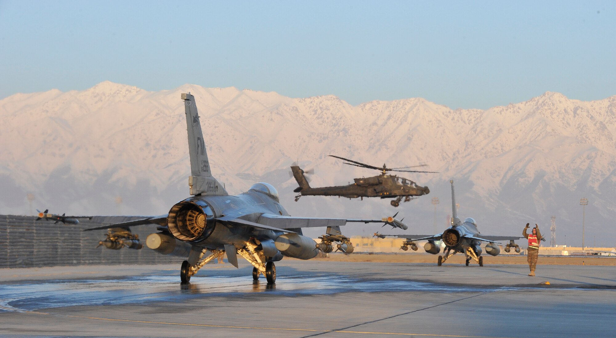 A pair of U.S. Air Force F-16 Fighting Falcons prepare to taxi out to the runway while a U.S. Army AH-64 Apache helicopter flies past at Bagram Airfield, Afghanistan, Feb. 11, 2014. Both types of aircraft are used to support ground operations in Afghanistan. (U.S. Air Force photo by Senior Master Sgt. Gary J. Rihn/Released)