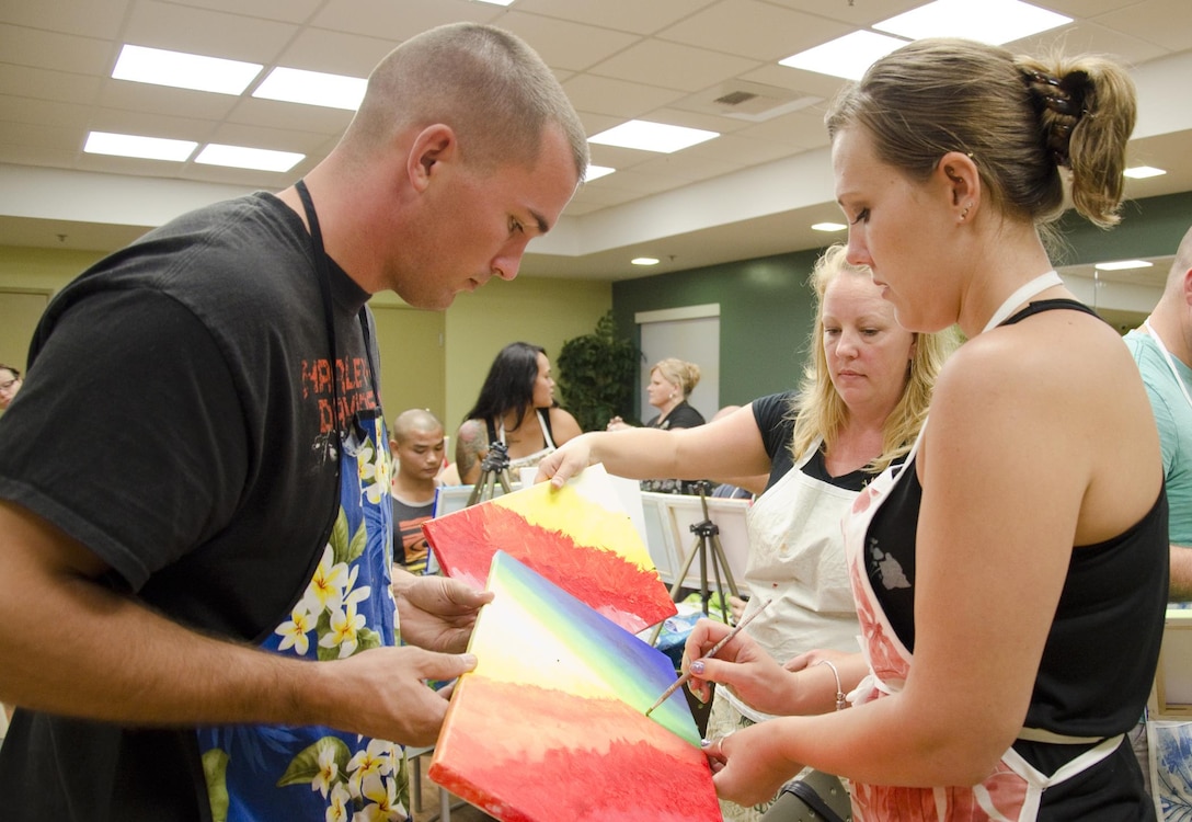 Sgt. William McGregor (left), an aviation ordnanceman with Marine Aviation Logistics Squadron 24, helps his wife, Julie (right), hold their paintings steady as she works to make them match. Painting instructor Katrina Marvin (center) guides them during the MALS-24 Wine and Canvas event at the Mololani Community Center, Feb. 13, 2014. (U.S. Marine Corps photo by Kristen Wong)
