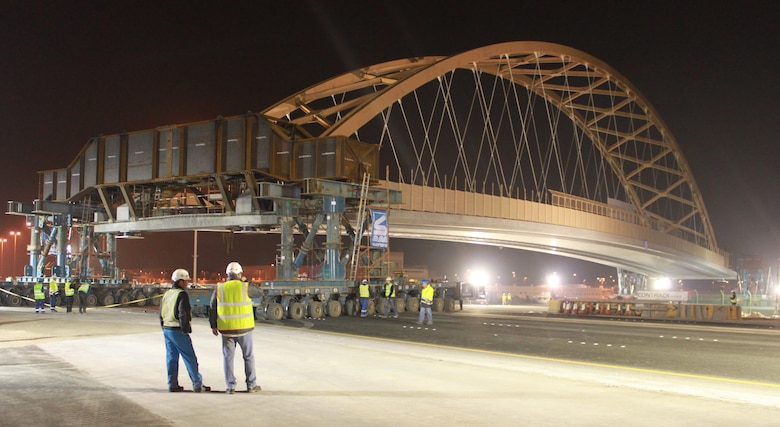 A tied arch bridge, known as the flyover bridge, was moved into place over the Khalifa Bin Salman Causeway connecting Naval Support Activity (NSA) Bahrain to the U.S. Navy port facility (NSA II). The 122.5 meter long, 21.4 meter high, 2650 metric ton bridge was constructed on NSA II, and will be driven across the causeway on self-propelled modular trailers (SPMT), and set in place on pre-constructed abutments. The greatest benefit of this process allows Bahrain traffic to be disrupted for hours as opposed to months it would have taken to build the bridge in place. The bridge move was a first of its kind for the Middle East District