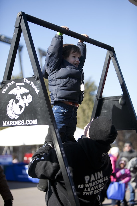 Staff Sgt. Thomas Hood, a member of the Enhanced Marketing Vehicle Team with the 8th Marine Corps District, helps a small child complete a pull-up at the Armed Forces Adventure Area in Fort Worth, Texas, Dec. 30, 2013. The 8th MCD Marines passed out prizes to anyone willing to complete some pull-ups and were available to answer any questions the public had about joining the Marine Corps. The AFAA was the official fan zone for spectators of the 2013 Bell Helicopter Armed Forces Bowl to interact with service members and their gear before, during and after the game.