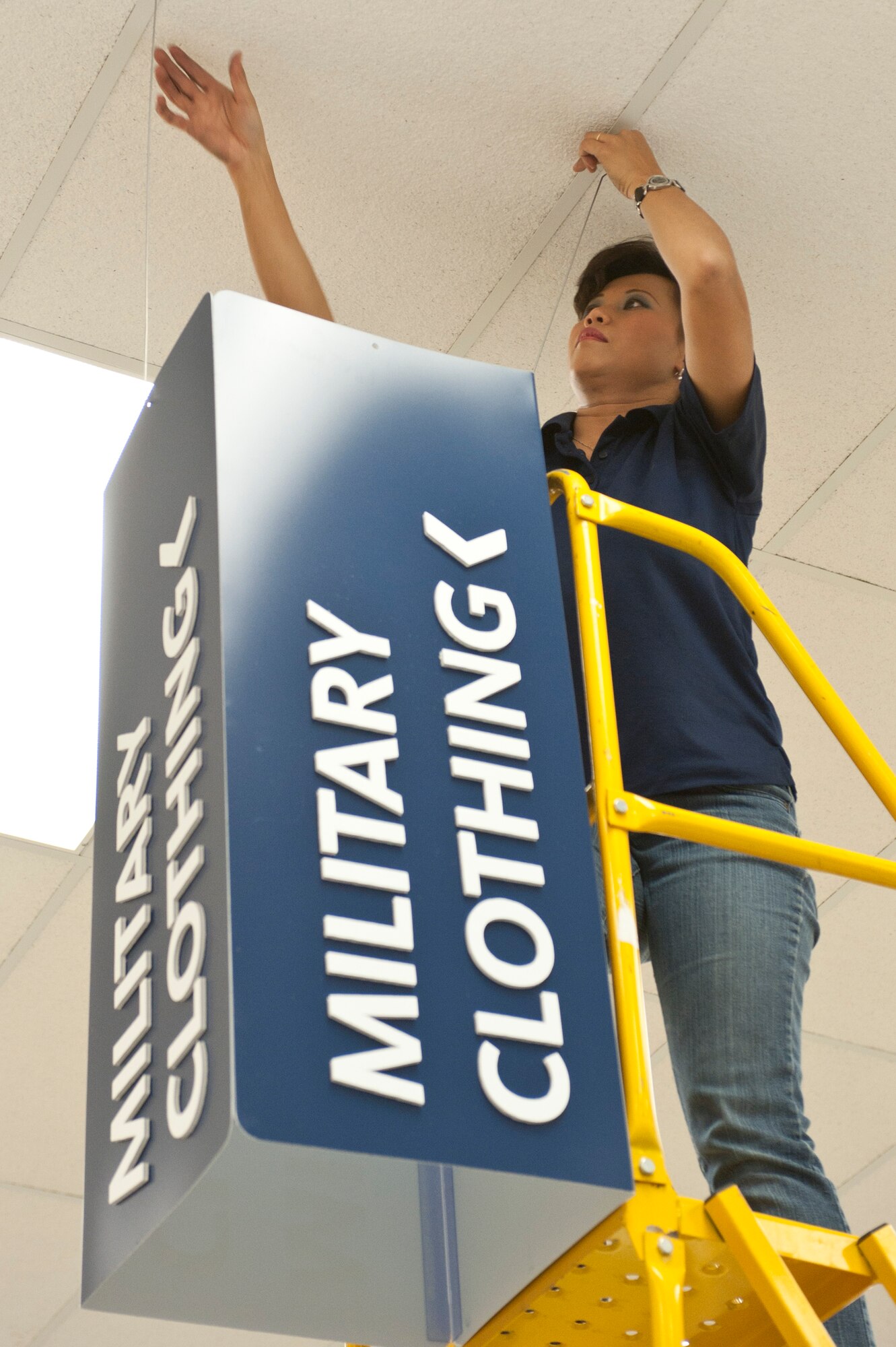 Susitian Jansen, an Exchange visual merchandiser at Nellis Air Force Base, Nev., hangs a military clothing sales sign in the Exchange during the move of military clothing sales Jan. 10, 2014. Military clothing sales is temporarily located in the outdoor living area in the Exchange right next to the Power Zone. (U.S. Air Force photo by Senior Airman Matthew Lancaster)