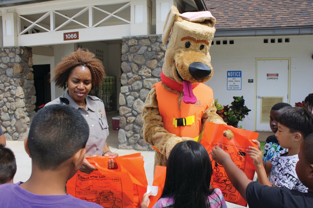Bobber the Water Safety Dog and Corps of Engineers Park Ranger Angela Jones pass out summer water safety packages to Fort Shafter Elementary School students during a water safety presentation at the Fort Shafter community center pool. 