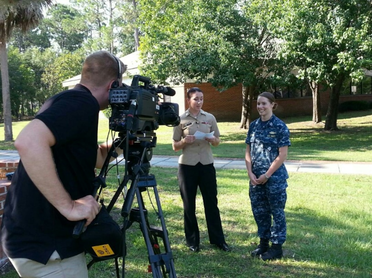 Two sailors stand next to each other outdoors while a videographer checks a camera on a tripod.