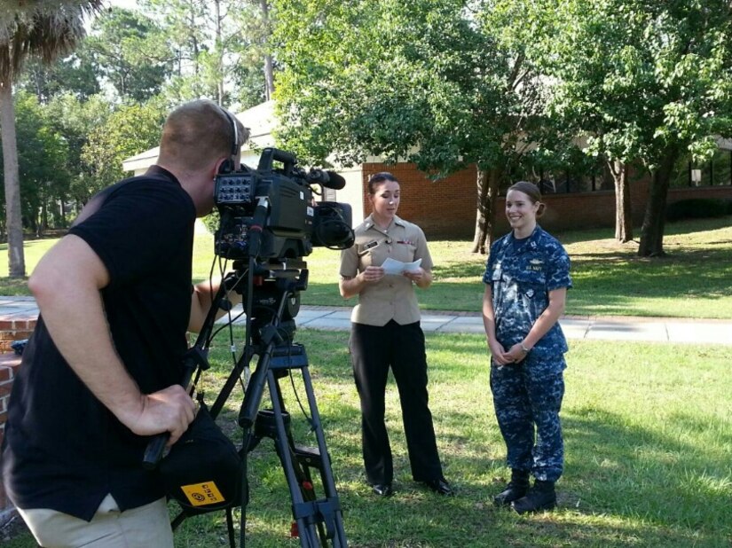 Two sailors stand next to each other outdoors while a videographer checks a camera on a tripod.