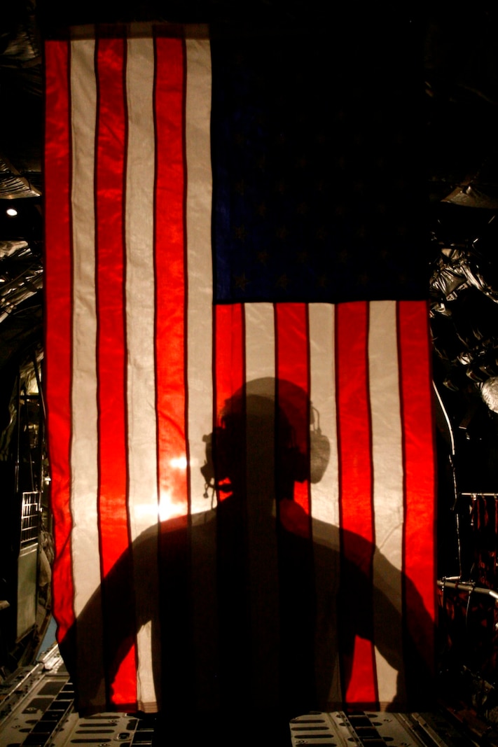 Corporal Jacob Hall, a crew chief with Marine Aerial Refueling Transportation Squadron 352, stands in front of an American Flag as a forklift shines its light, casting his shadow behind him after a battlefield illumination mission aboard Camp Bastion, Afghanistan, July 18, 2014. Battlefield illumination missions are implemented to light up areas in support of nighttime coalition operations within Regional Command (Southwest). (U.S. Marine Corps Photo by: Sgt. Frances Johnson/Released)