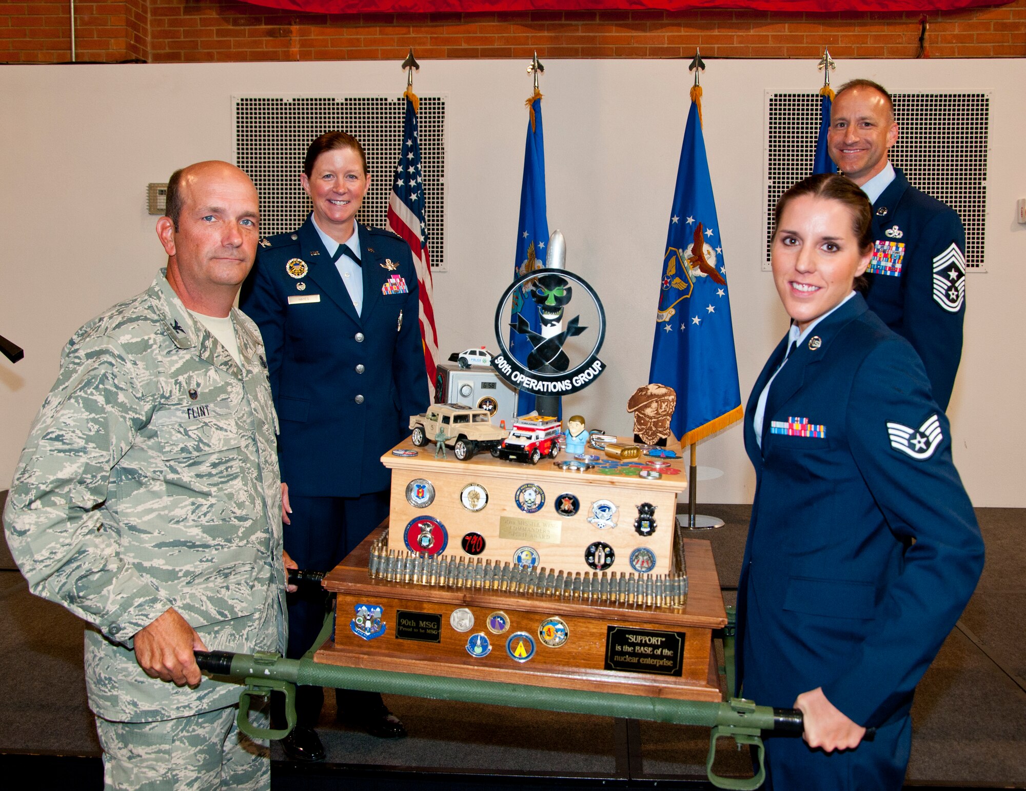 Col. Trevor Flint, 90th Maintenance Group commander, and Staff Sgt. Katie Coble, 90th Maintenance Operations Squadron Facilities Maintenance Team instructor, hold the 90th Missile Wing Spirit Award and pose with Col. Tracey Hayes, 90th Missile Wing commander, and Chief Master Sgt. Samuel Couch, 90th MW command chief, July 29, 2014, during the 90th MW Second Quarter Awards Breakfast in the Fall Hall Community Center. The spirit award is given to the group that shows the most exuberance and has the loudest chants at the award ceremony, and the 90th MXG won. (U.S. Air Force photo by Airman 1st Class Jason Wiese)