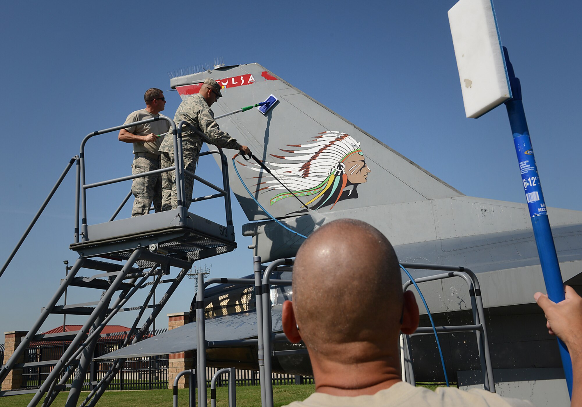 Chief Master Sergeant Dennis Dipiazzo, 138th Communications Flight Chief, stands ready to scrub as Lieutenant Colonel Travis Brown, 138th Maintenance Squadron Commander and Chief Master Sergeant Tracey Weaver, 138th Aircraft Maintenance Chief, power wash the tail of an F-16 static display aircraft at the Tulsa Air National Guard base, Tulsa Okla., 29 July 2014.  Members of the 138th FW Chiefs Council gathered to wash the static aircraft in an effort to keep them looking nice for public display.  (U.S. National Guard photo by Senior Master Sgt.  Preston L. Chasteen/Released)