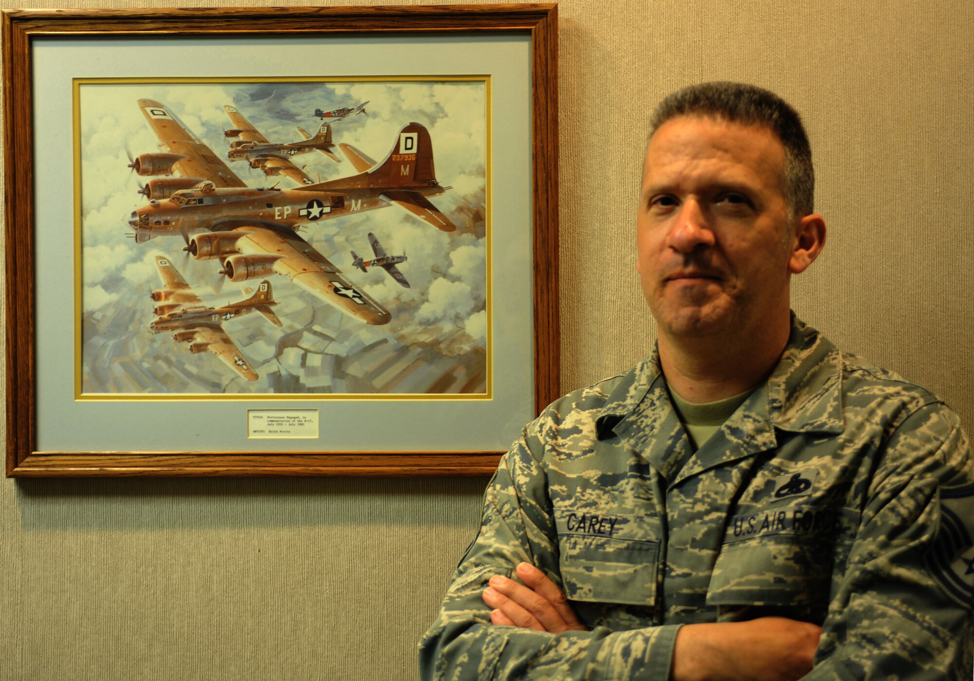 Master Sgt. Matthew Carey, 28th Bomb Wing Treaty Compliance Office superintendent, stands by an illustration of several B-17 Flying Fortress bombers on a mission during World War II displayed in the hallway outside his office. Carey said the image reminds him of his grandfather, B-17 tail gunner Staff Sgt. James “Rae” Carey, and his experiences during World War II after he signed in for duty at a base in England one day after the invasion of Normandy began.  (U.S. Air Force photo by Senior Airman Yash Rojas/Released)