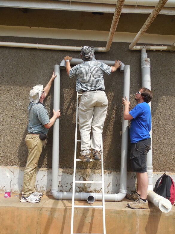 Jim Goedert, EWB-NE Professional; Dwight Hanson EWB-NE-P President ; and Ben Pavlik EWB-NE Student install the modified first flush piping. on St. John’s Clinic. The first flush system is a vertical pipe into which the first rainfall from the roof washes at the beginning of a rainfall event. The water from the first flush contains dirt and debris that accumulates on the roof during the dry period. Once the vertical pipe of the first flush system fills; cleaner water flows over it and into the water tanks. The first flush system helps prevent the tanks from filling with dirt and keeps the water in the tank cleaner.