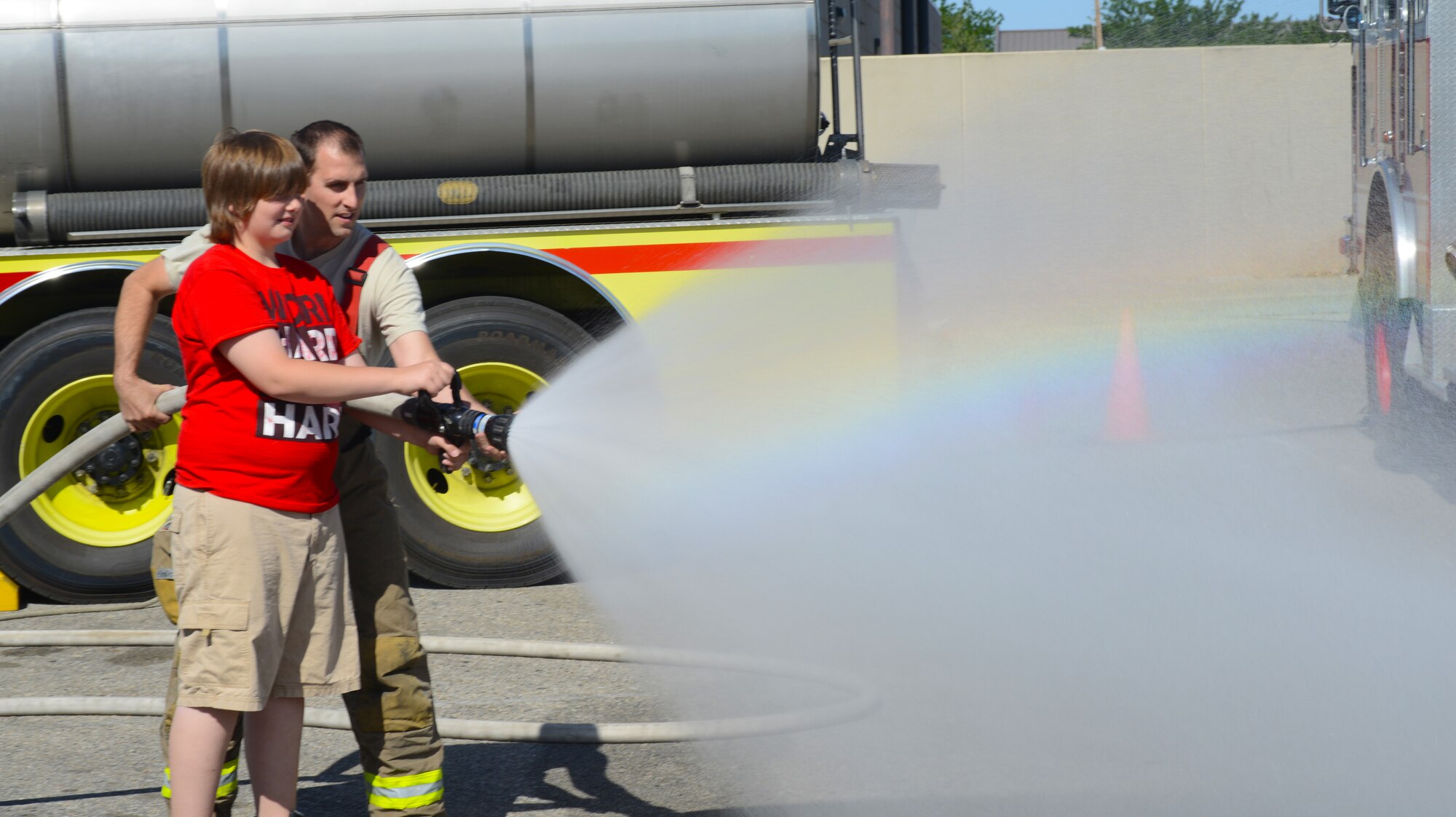 Staff Sgt.Travis Hire, 366th Civil Engineering Squadron NCO in charge of the emergency control center, assists Alec Parks, son of Master Sgt. Eric Parks assigned to the 366th Security Forces Squadron Alpha flight chief, with spraying the fire hose June 6, 2014, at Mountain Home Air Force Base, Idaho. Mountain Home AFB held a safety fair in the parking lot of the base exchange in an effort to highlight specific safety concerns during the 101 Critical Days of Summer. (U.S. Air Force photo by Senior Airman Benjamin Sutton/Released)