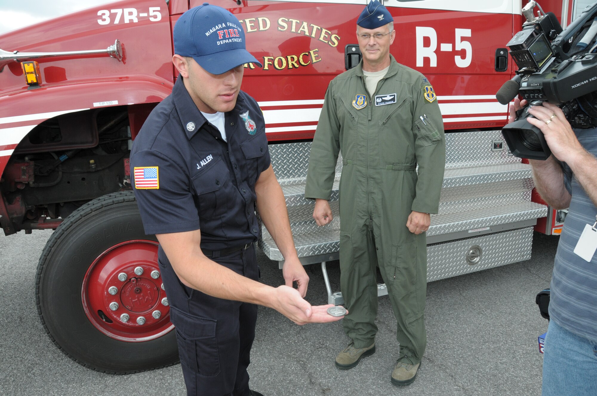 Joseph Allen, 914th Fire Emergency Services Department of Defense civilian and Air Force Reserve Firefighter/Emergency Medical Technician, shows local media  the commander’s coin presented to him by Col. Steven Parker (center), 914th Airlift Wing Commander, at the Niagara Falls Air Reserve Station, N.Y. June 12, 2014. The coin was presented to Allen for responding and treating a woman involved in a serious auto accident. (U.S. Air Force Photo by Peter Borys) 