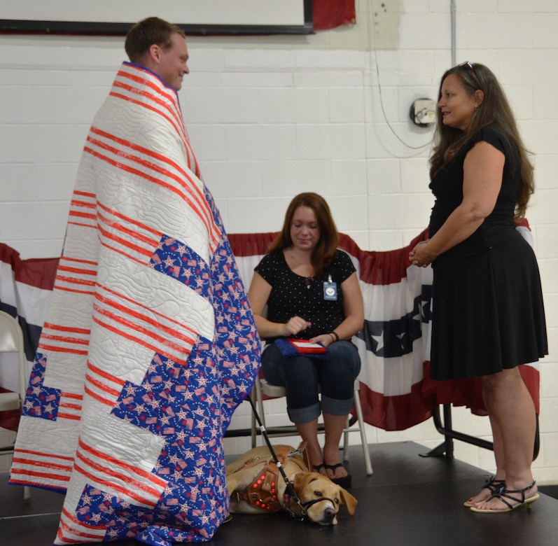 Anne Mixon from Quilts of Valor (right), presents retired U.S. Marine Sgt. Kyle Miller, a quilt in recognition of his military service while Miller’s wife Vanessa and his new canine companion Ava, look on June 19, 2014, at the Naval Consolidated Brig Charleston. The Quilts of Valor Foundation has a goal to cover all combat service members and veterans touch by war with comforting and healing Quilts of Valor. (U.S. Air Force photo/Eric Sesit) 