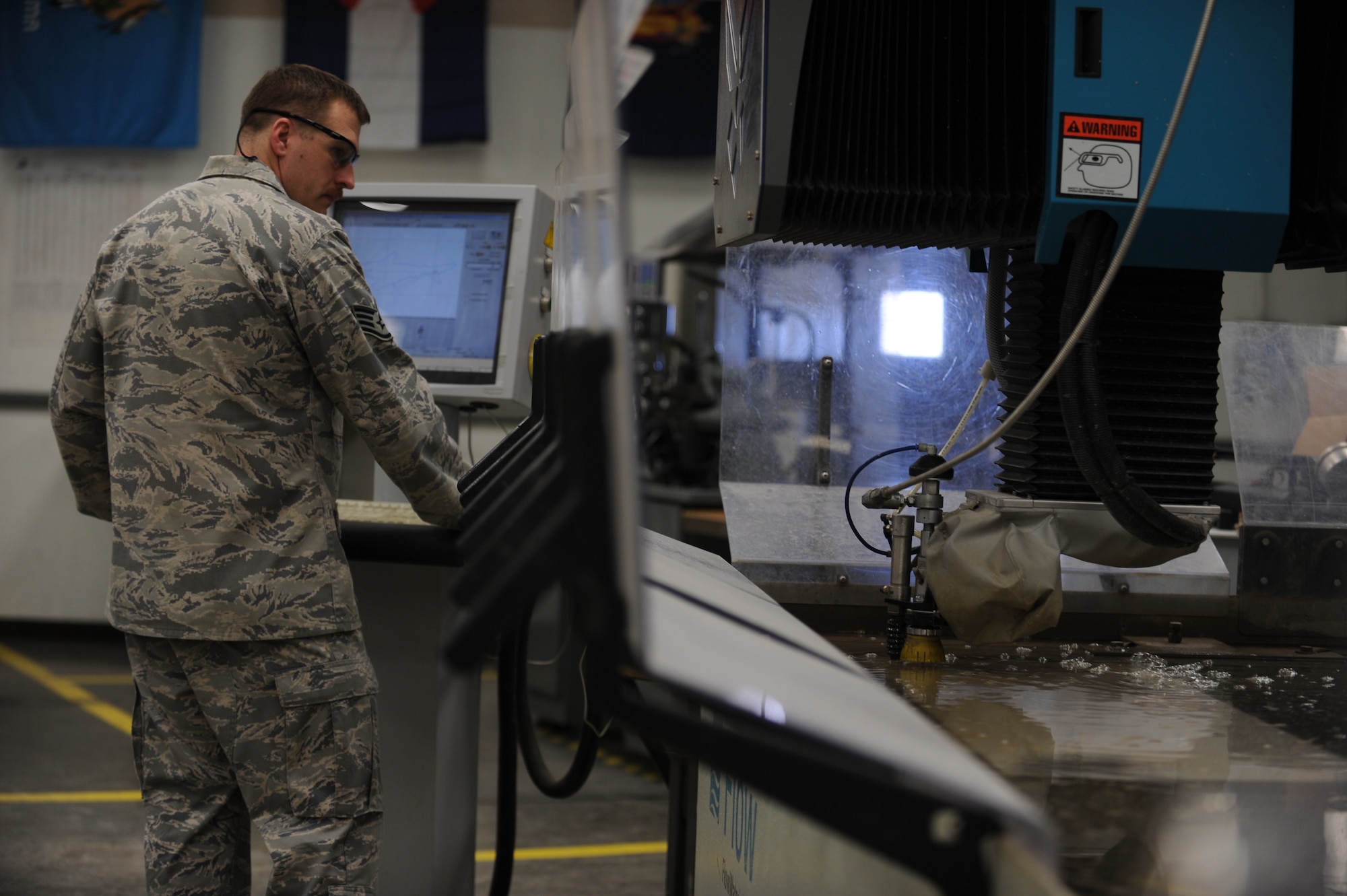 Staff Sgt. Kenny Gayoso, a 19th Equipment Maintenance Squadron duty title, observes a flow water jet abrasive cutter work March 11, 2014, at Little Rock Air Force Base, Ark. The cutter is commonly connected to a high-pressure, water pump where the water is then ejected from the nozzle, cutting through the material by spraying it with the jet of high-speed water. (U.S. Air Force photo by Airman 1st Class Cliffton Dolezal)